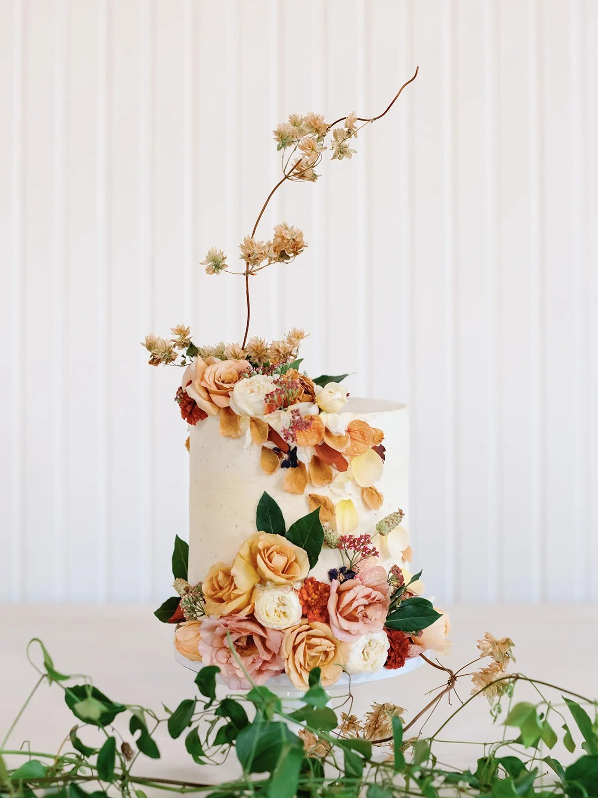 A tall, white, multi-tiered wedding cake decorated with fresh pink, cream, and orange flowers and greenery, set on a table with green vine decorations against a light background.