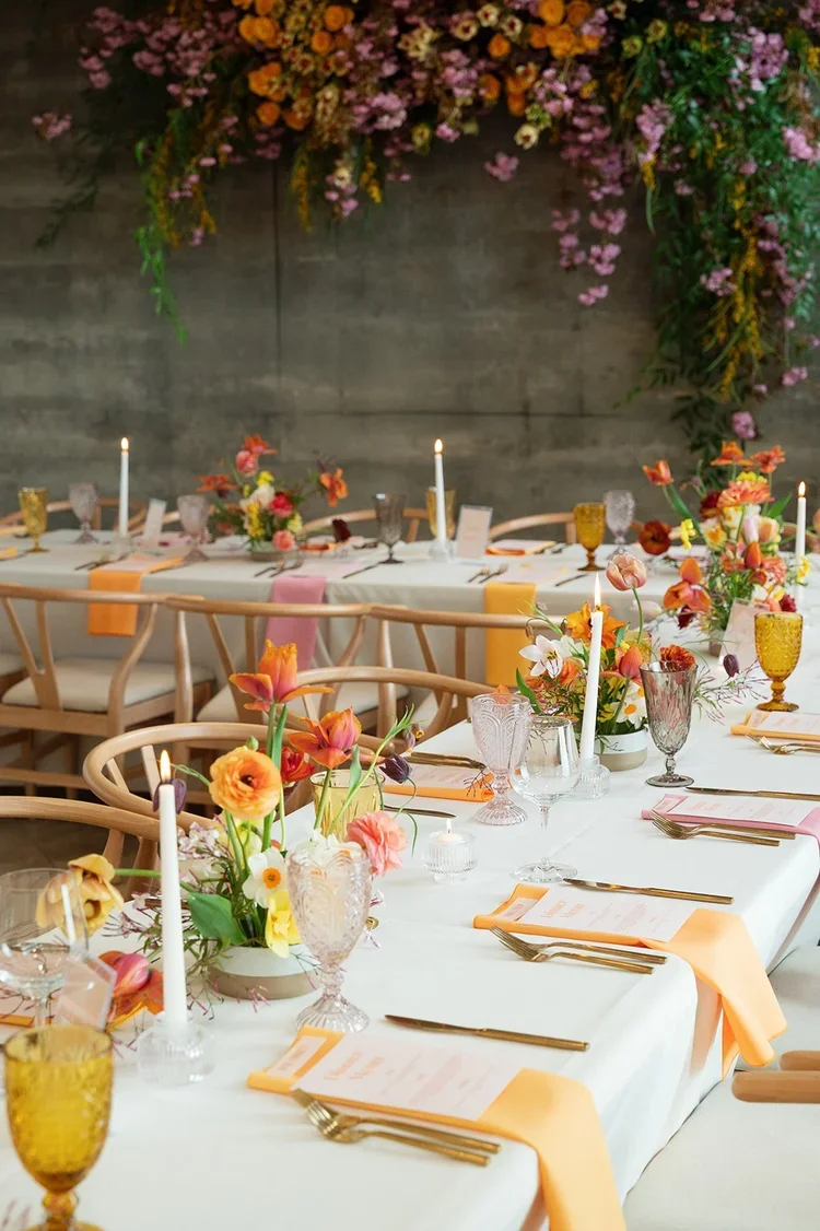 Wedding or event table decorated with pink, orange, and white flowers, candles, and elegant glassware against a concrete wall with a floral arrangement overhead.