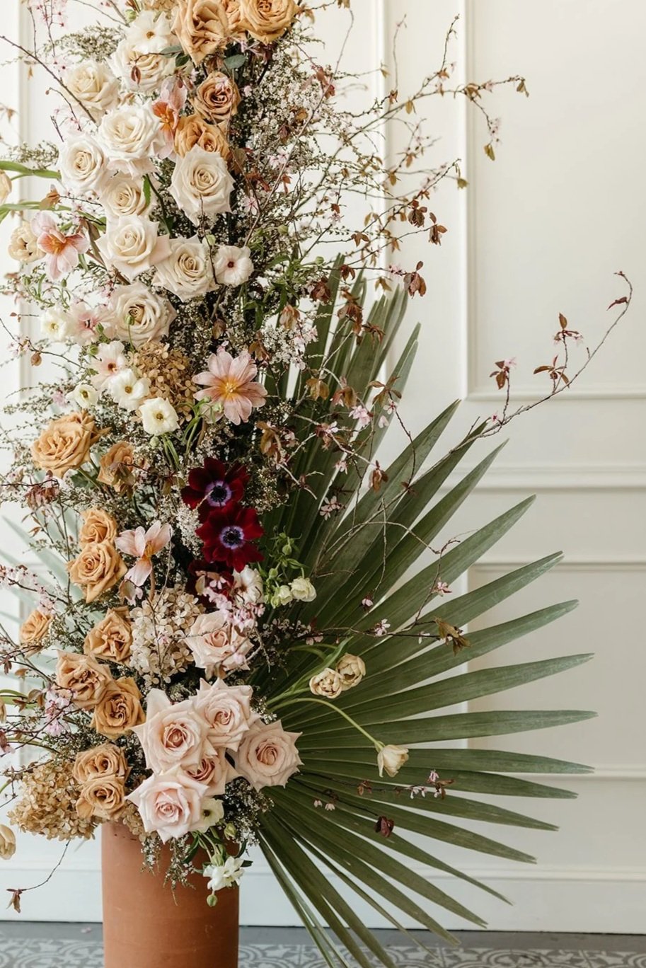 A large floral arrangement in a brown vase with white, cream, pink, and deep red flowers, including roses, anemones, and other mixed blossoms, accented by tall green leaves and wispy branches, placed against a white paneled wall.