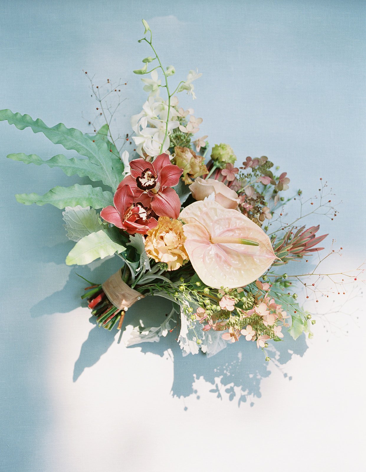 A colorful bouquet of various flowers including orchids, anthuriums, and greenery on a pale blue background.