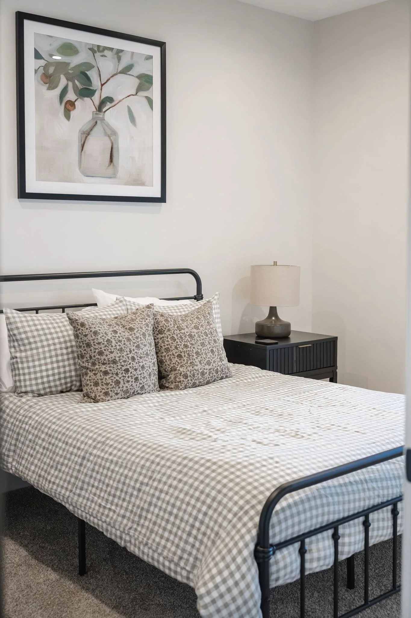 A bedroom with a black metal bed frame, gingham patterned bedspread, multiple leopard print pillows, and a nightstand with a beige lamp. A framed painting of a vase with green leaves and small fruits hangs above the bed.