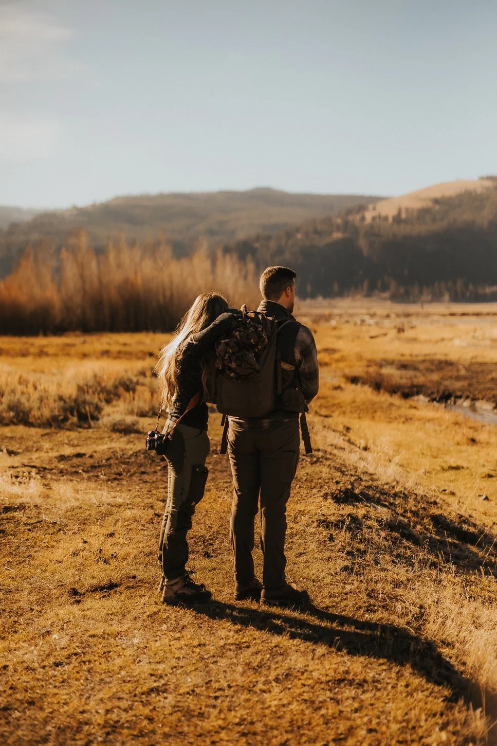 A man and woman with backpacks standing outdoors in a field during sunset, looking at distant hills.