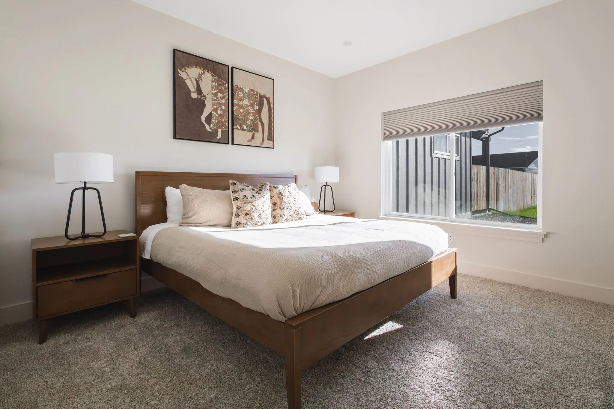 A bedroom with a wooden bed frame, white bedding, and pillows with patterned covers. On either side of the bed are matching black tripod lamps. Above the bed are two art pieces featuring a horse. There is a window with blinds to the right, showing a 