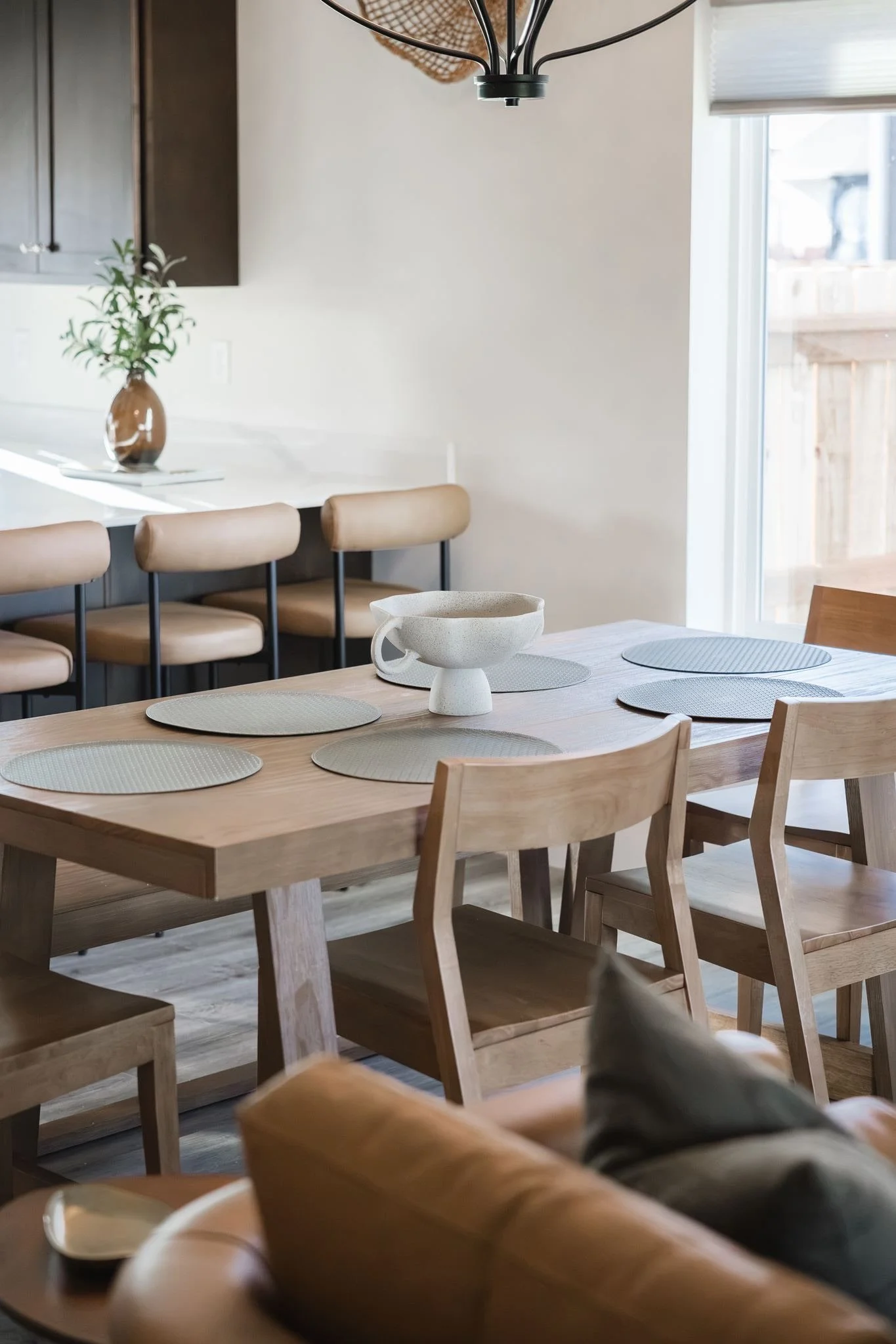 A dining area with a wooden table, six chairs, and placemats. A bowl or vase is in the center of the table. In the background, there are beige chairs along a white countertop with a potted plant, dark cabinets, and a window with a blind letting in na