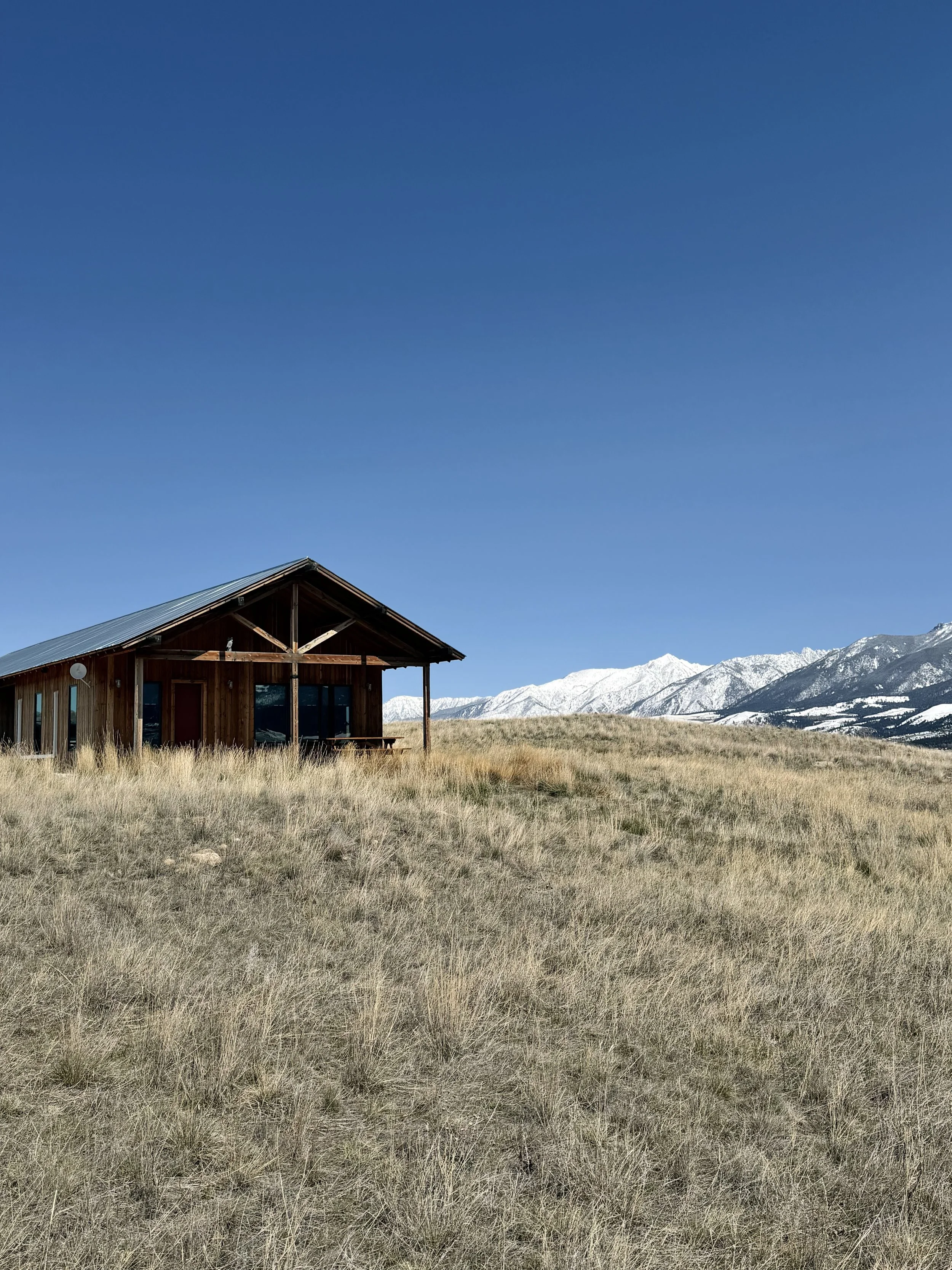 A wooden house or cabin with a metal roof in a grassy field, snow-capped mountains in the background, and a clear blue sky overhead.