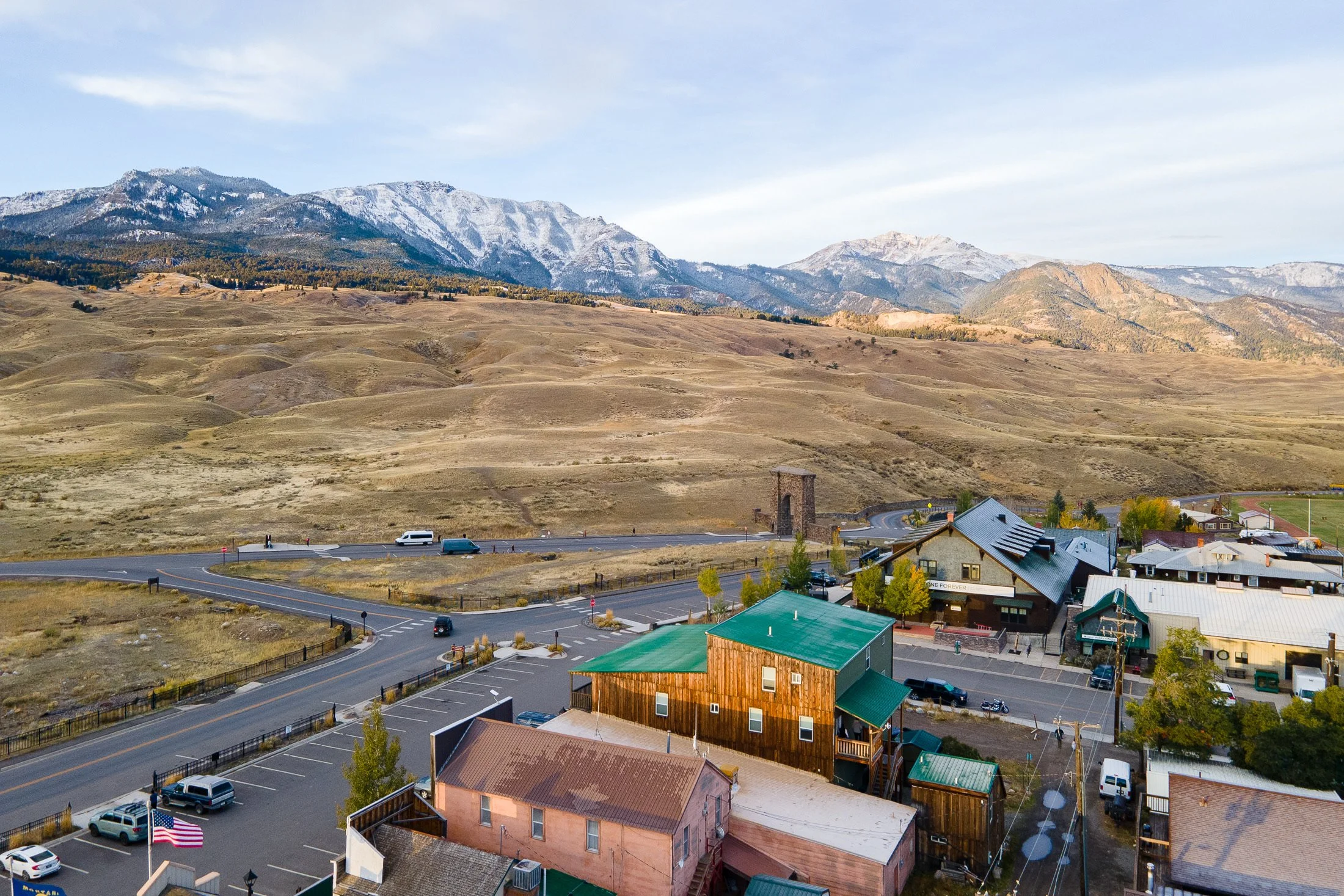 A scenic view of a town with colorful buildings, parking lots, and streets at the base of a mountain range with snow-capped peaks under a blue sky.