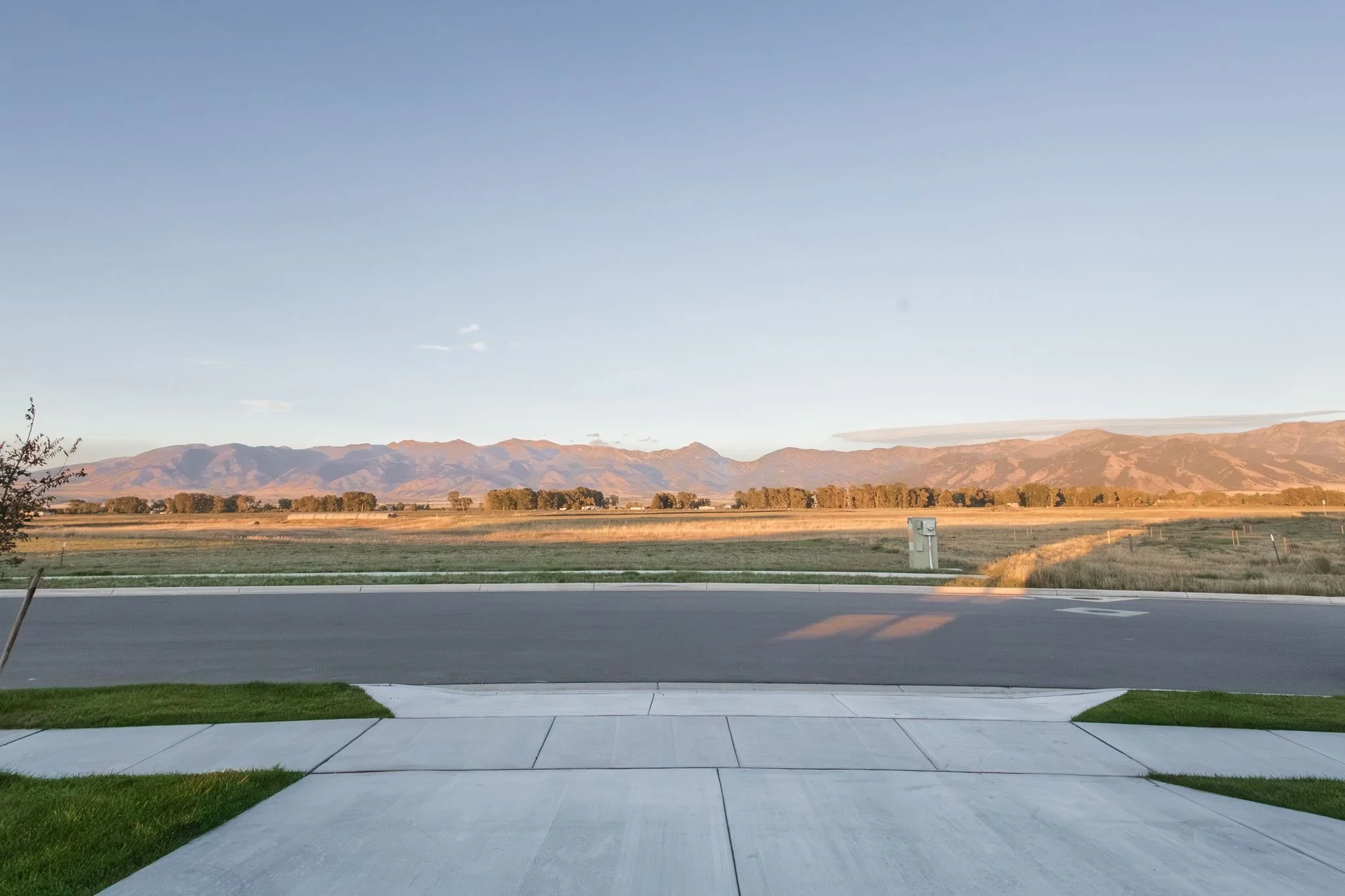 View of a suburban street with a sidewalk and lawn in the foreground, a barbed-wire fence and open field with trees, and mountains in the distance under a clear blue sky.