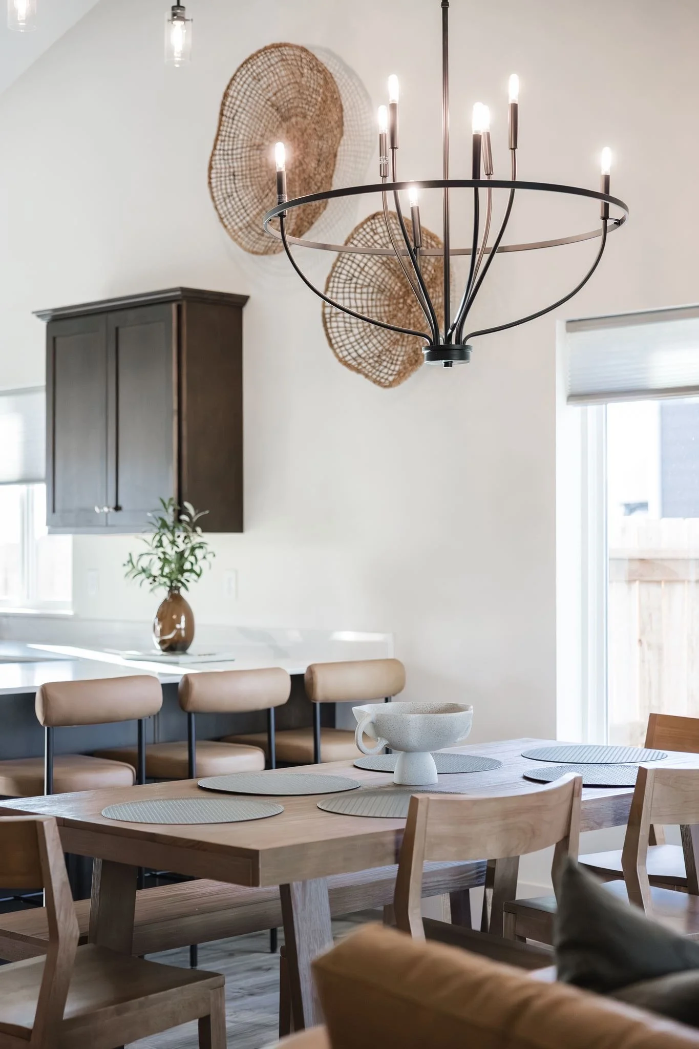 Modern dining room with a wooden table, beige chairs, a white centerpiece, a black chandelier, a wall with woven art, a dark cabinet, and windows with blinds.