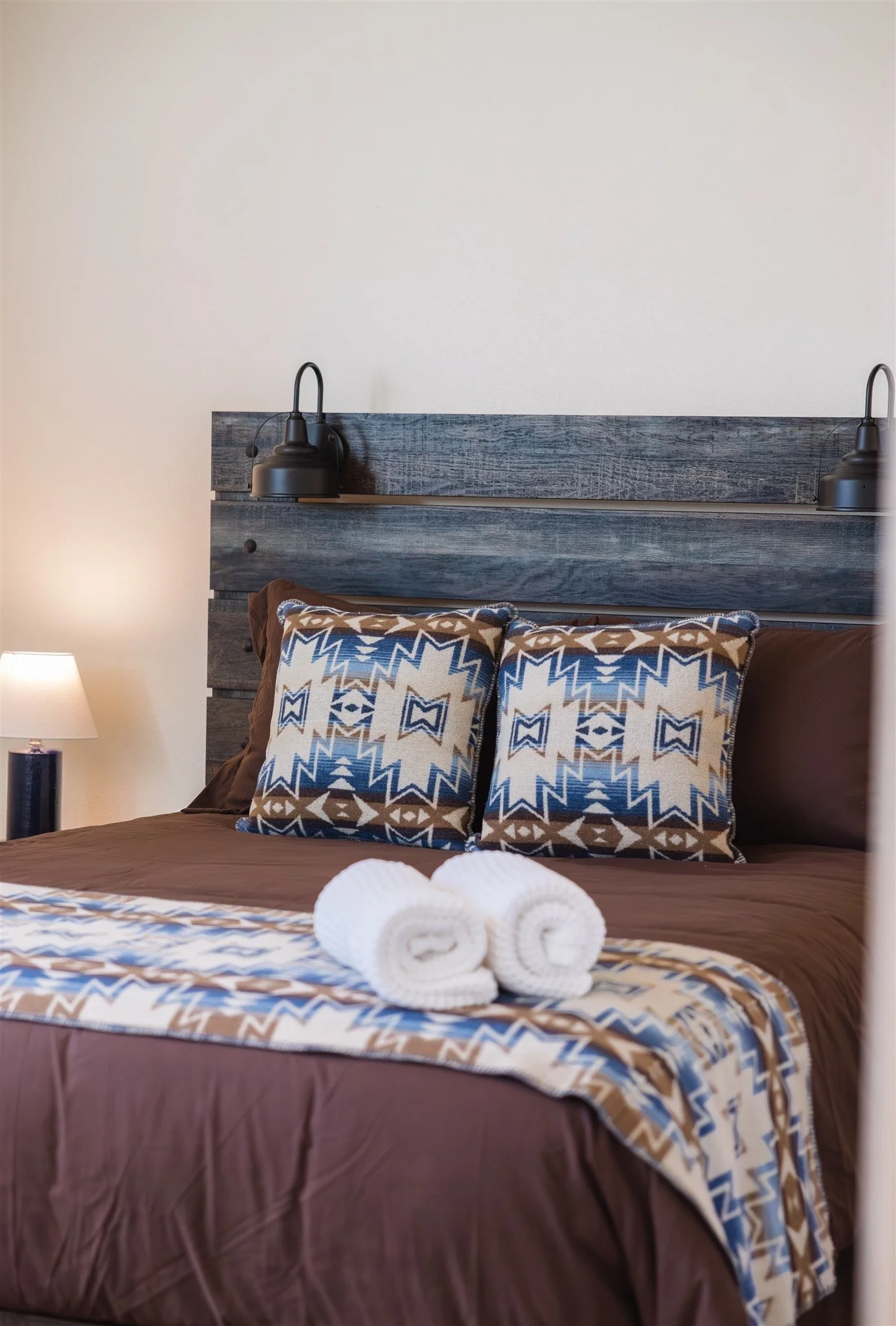 A neatly made bed with brown sheets and a decorative quilt, two decorative pillows with geometric patterns in blue, brown, and white, three rolled white towels on the bed, a wooden headboard with two black wall-mounted lamps on either side, a partly 