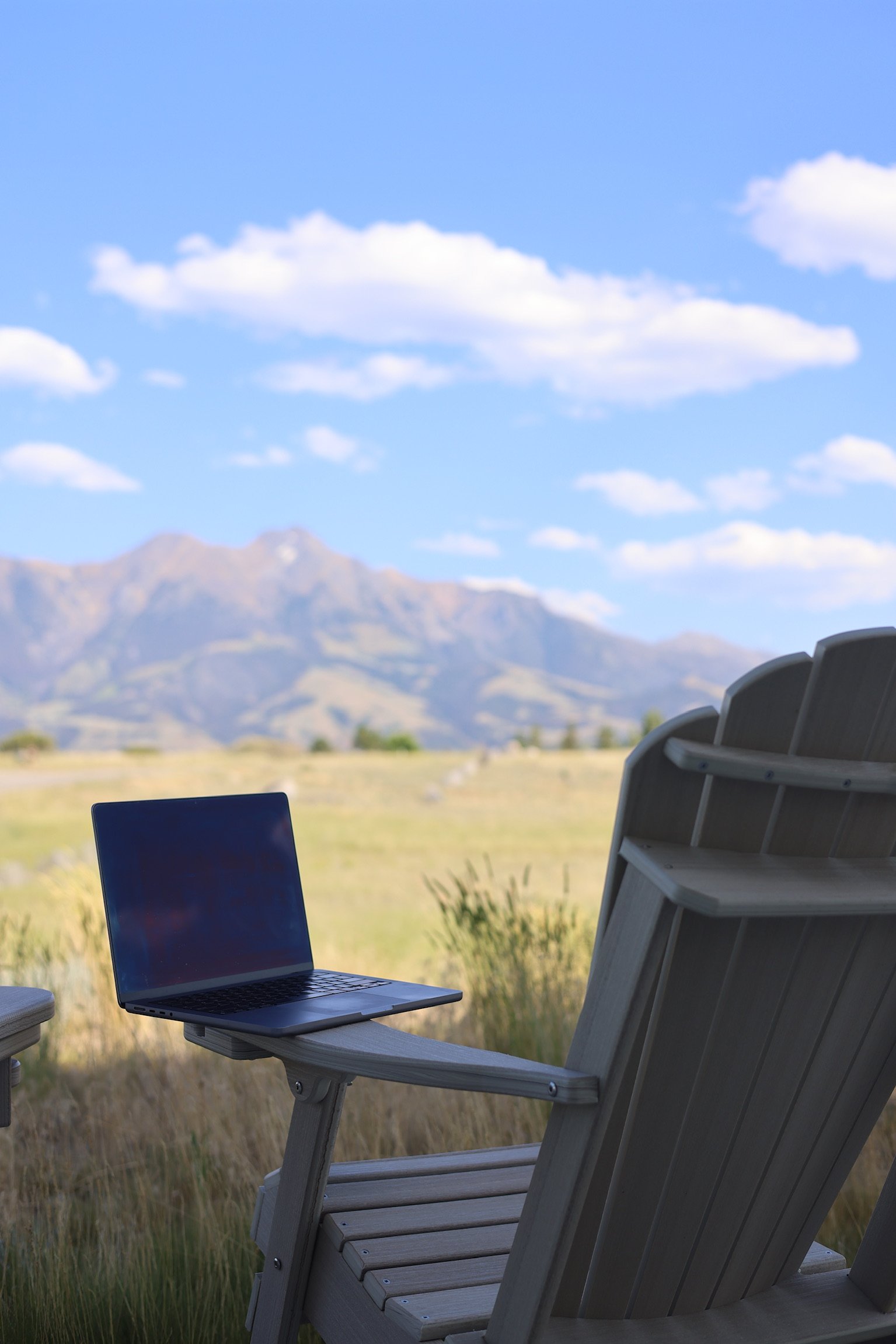 A laptop on an Adirondack chair outdoors with mountains and a blue sky with clouds in the background.