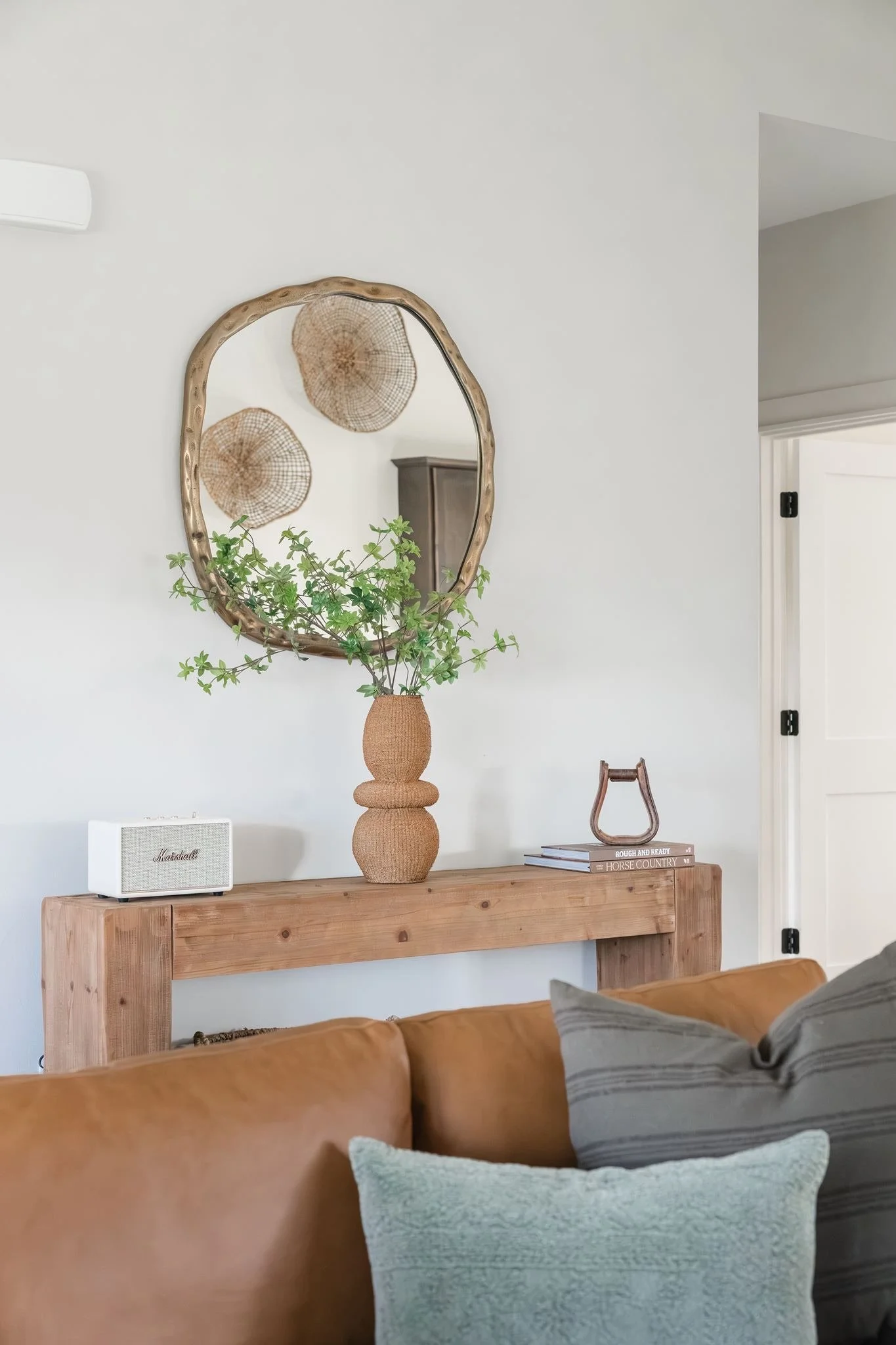 Living room with a wooden console table decorated with a textured vase and a leafy plant. A round mirror with a wooden frame hangs on the white wall above the table, reflecting two woven wall hangings. A white speaker is on the table, and part of a l