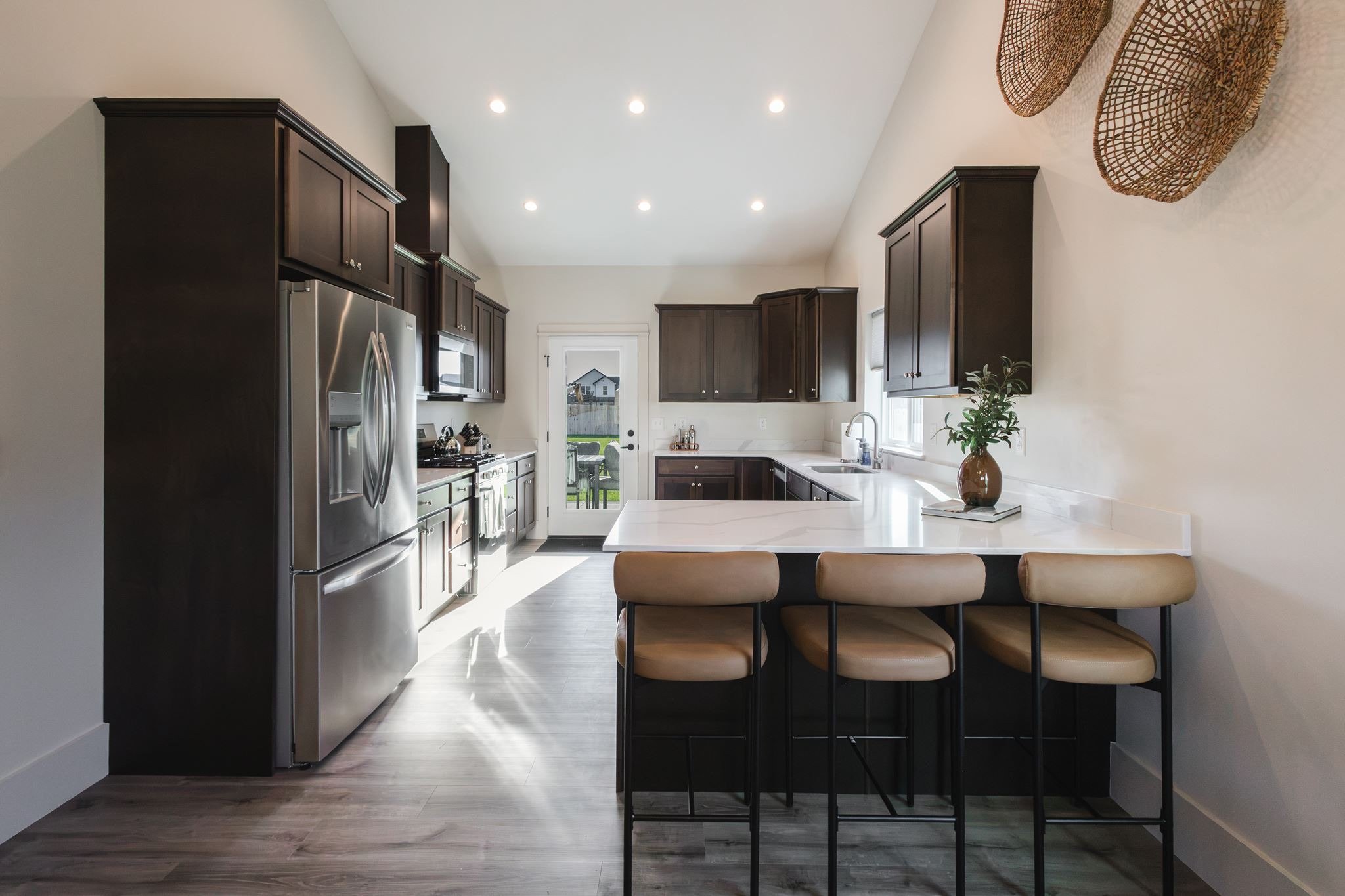 Modern kitchen with dark cabinetry, stainless steel appliances, white countertops, and a breakfast bar with beige chairs. Pots and a plant are visible on the counter, with a glass door leading outside.