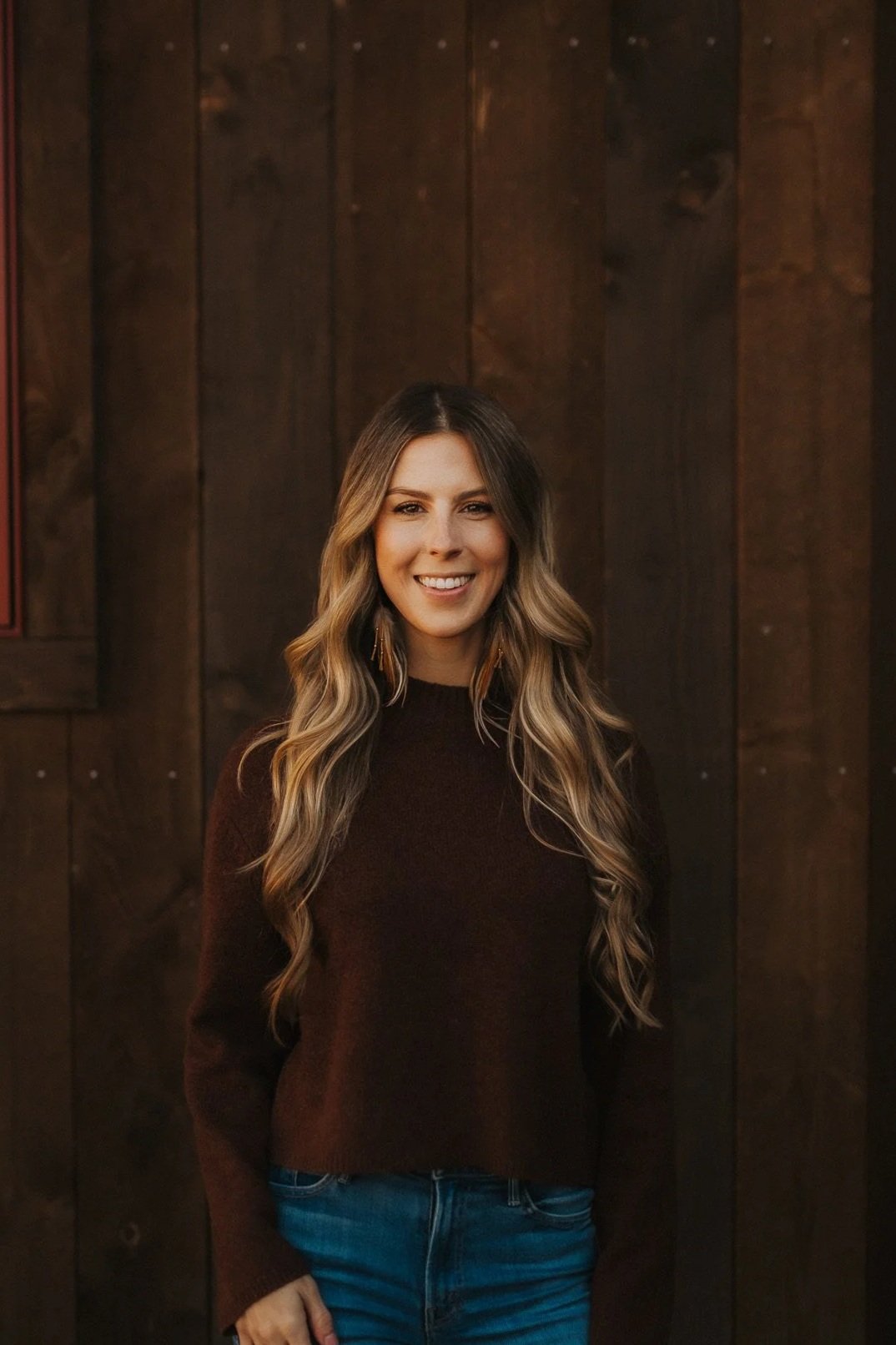 A woman with long blonde hair, wearing a black sweater and a necklace, smiling in front of a light-colored wall and some green foliage.