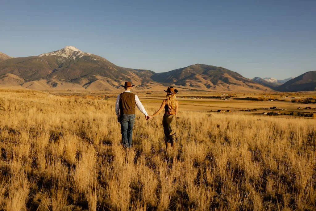 A couple holding hands walking in a vast grassy field with mountains in the background during sunset.