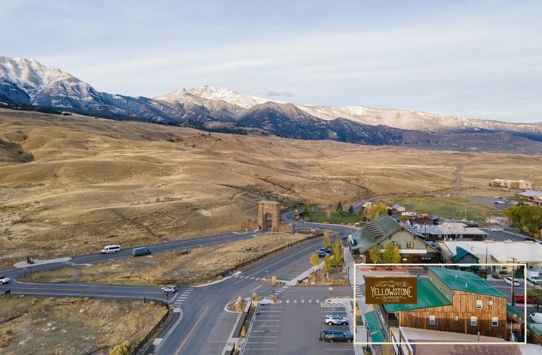 A small town called Yellowstone Outpost with buildings, parking lot, and a road, situated at the base of snow-capped mountains.