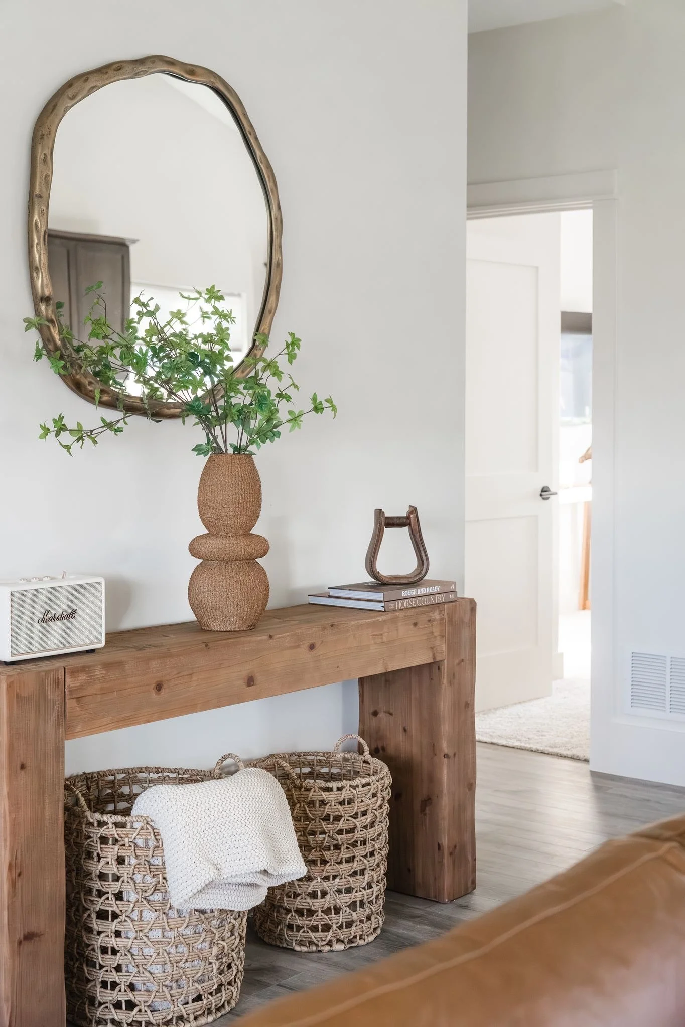 A wooden console table with a woven wicker basket and a white pillow inside it. On top of the table, there are two stacked books, a decorative wooden or metal object, a small white speaker, and a green plant in a textured vase. A mirror with a wavy, 