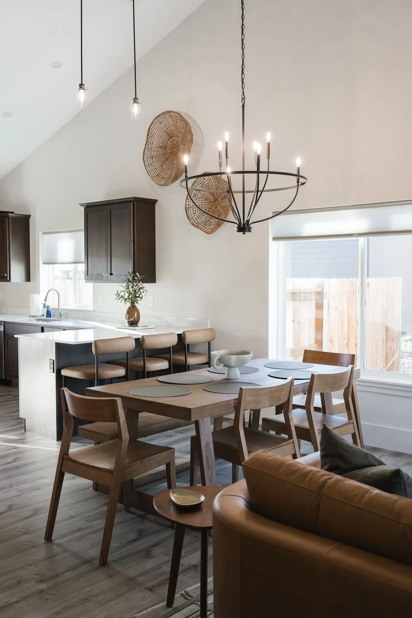 Modern kitchen and dining area with a wooden dining table, chairs, a chandelier, pendant lights, kitchen cabinets, and a large window with blinds.