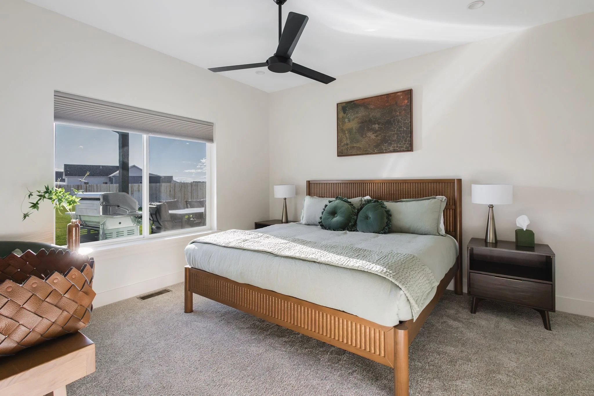 A bedroom with a wooden bed frame, white bedding, green pillows, two nightstands with lamps, a painting above the bed, a window with a view of a backyard with a grill, and a ceiling fan.