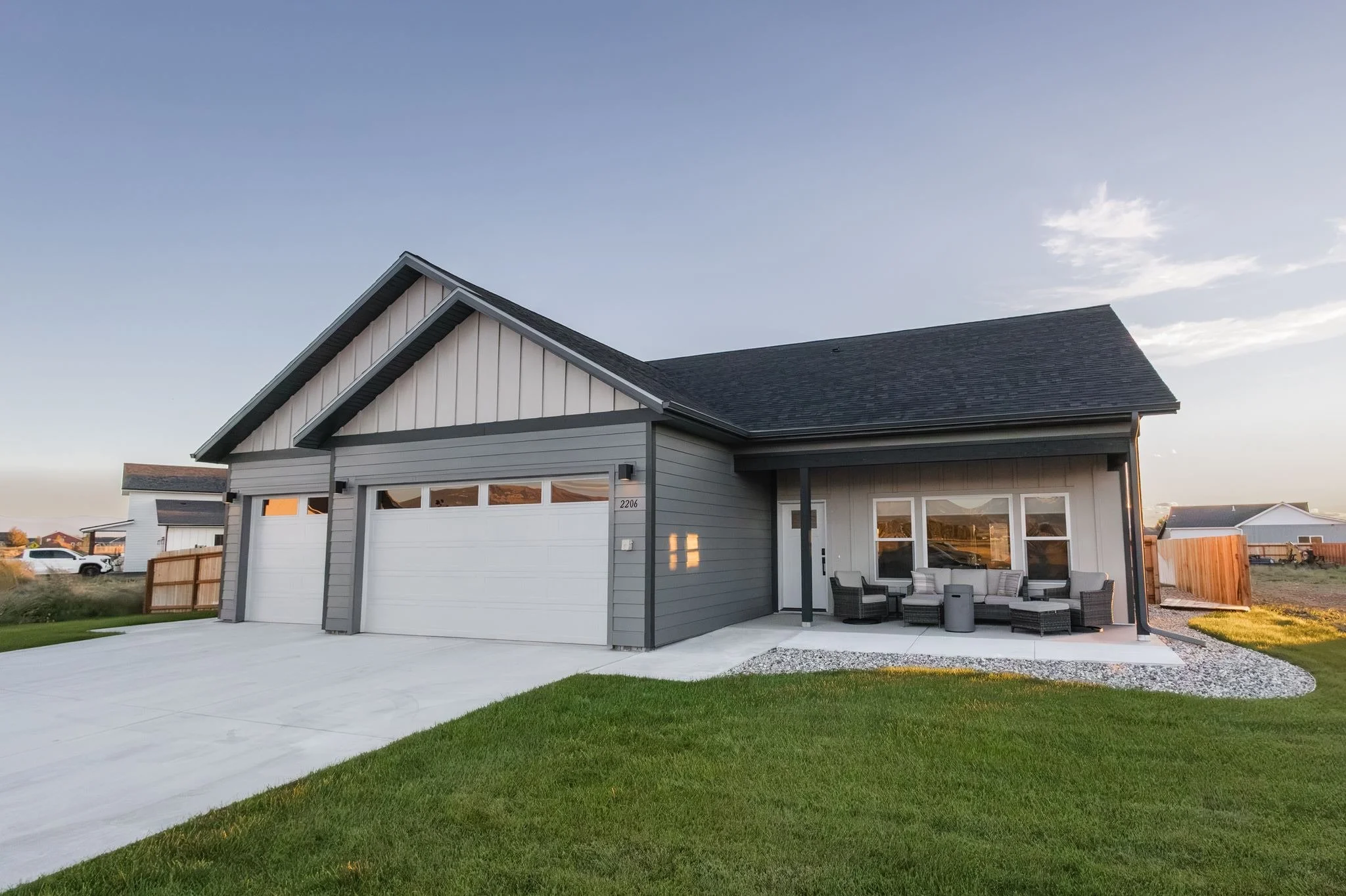 A modern house with a gray exterior, white garage door, and a small outdoor seating area with chairs and a table, set on a well-maintained lawn with neighboring houses in the background under a partly cloudy sky.