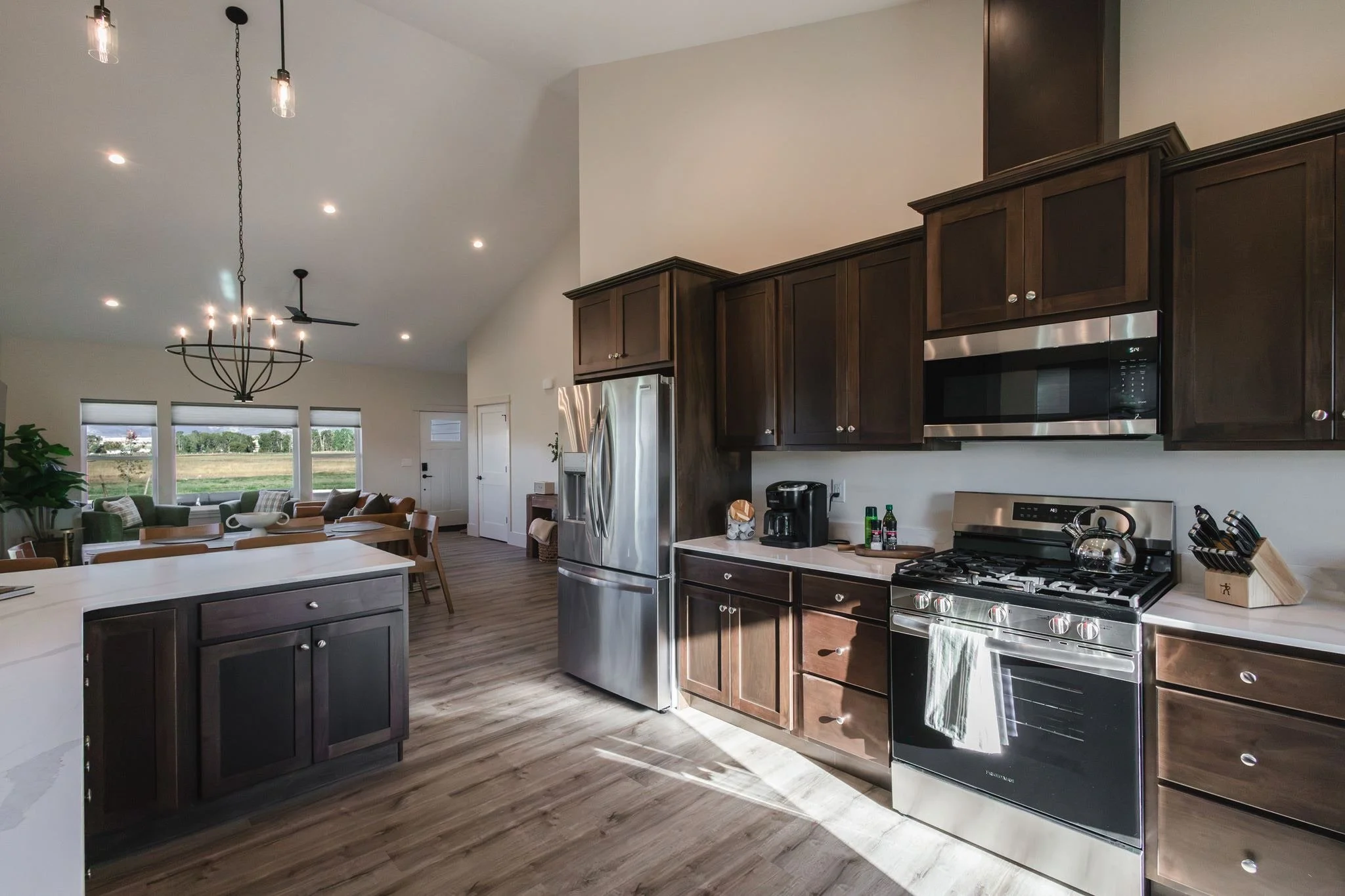 Kitchen with dark wood cabinets, stainless steel appliances, and a white countertop. Open living space with large windows, a dining table, and a living area with couches.
