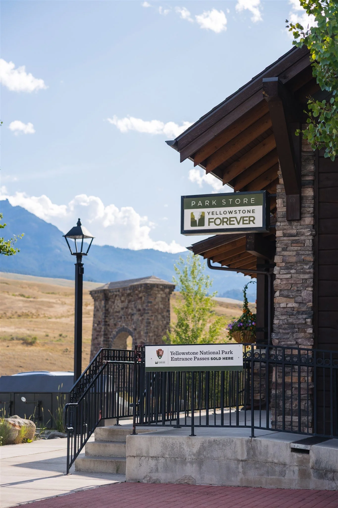 Entrance of Yellowstone National Park with signs indicating park store and ticket sales. Stone archway and lamp post are visible, with mountainous landscape in the background under a clear sky.