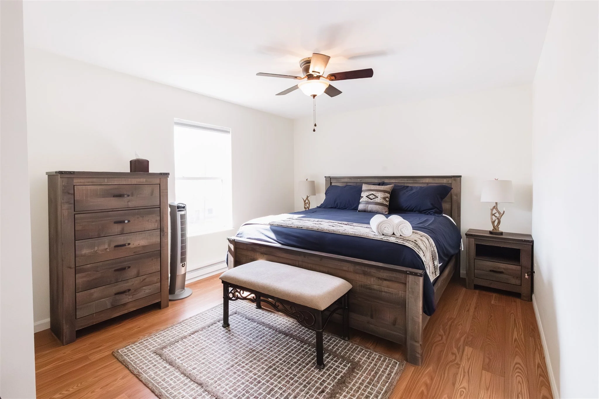 A bedroom with a wooden bed, navy blue bedding, and decorative pillows. There's a wooden dresser, a white table lamp on a side table, a window with blinds, a small bench at the foot of the bed, a rug, a tower fan, and a ceiling fan with lights.