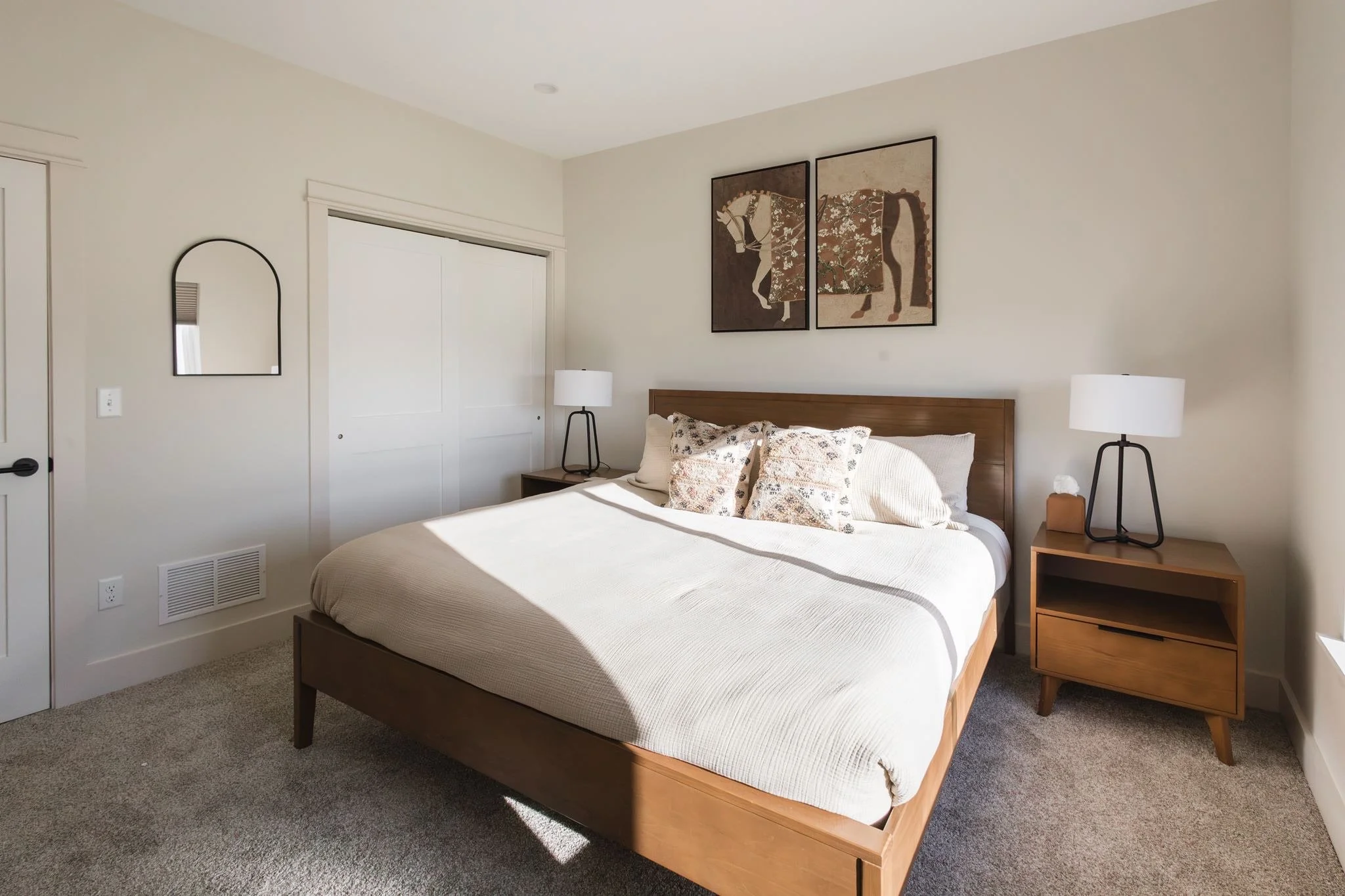 A bedroom with a wooden bed frame and a white mattress, decorated with pillows. Two matching black table lamps sit on wooden nightstands on either side of the bed. There is a large framed horse artwork on the wall above the bed, and a small arched mi
