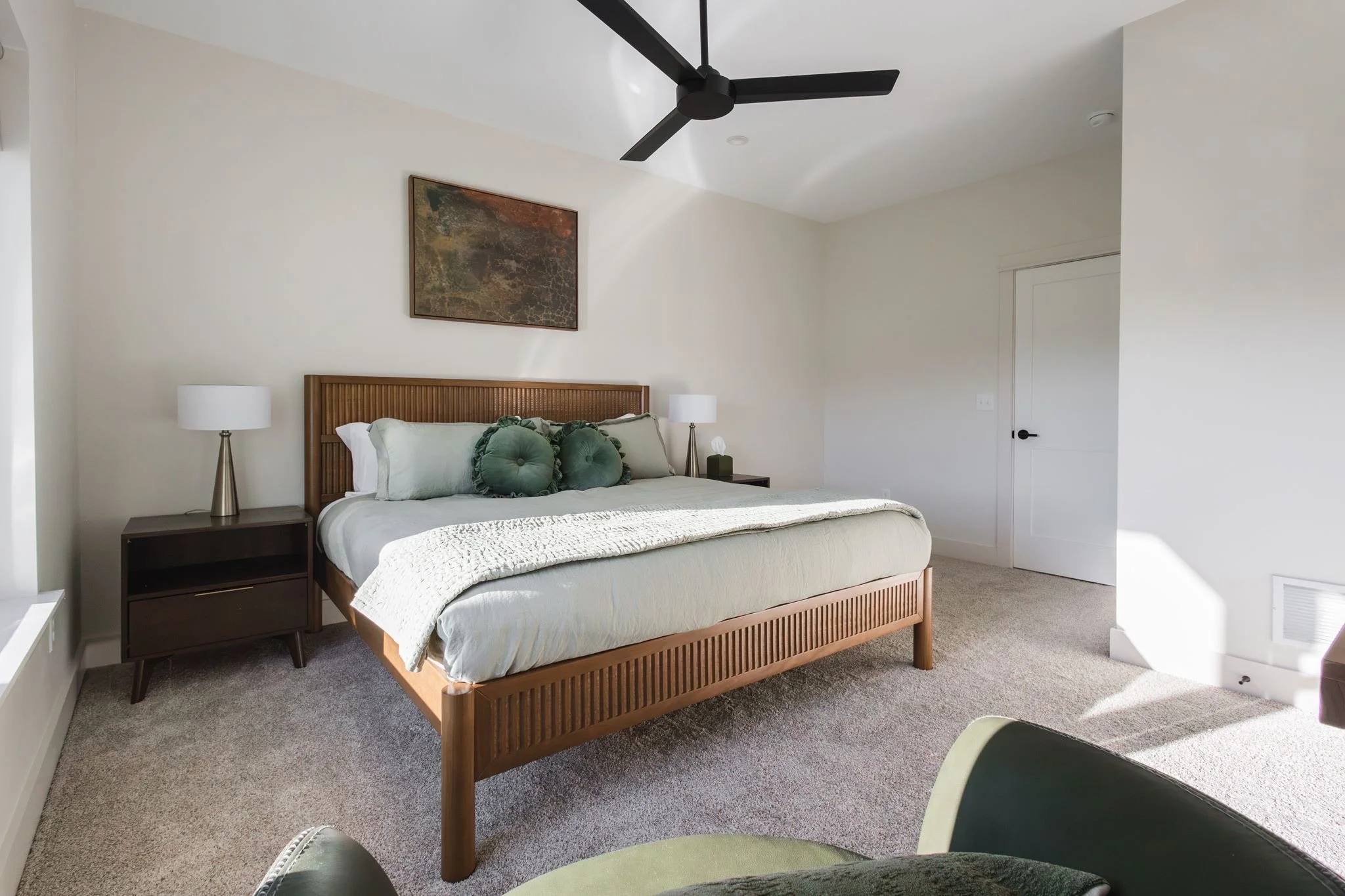 A bedroom with a wooden bed frame, white bedding, green pillows, two white lamps on nightstands, a rectangular wall art, a black ceiling fan, and a white door.
