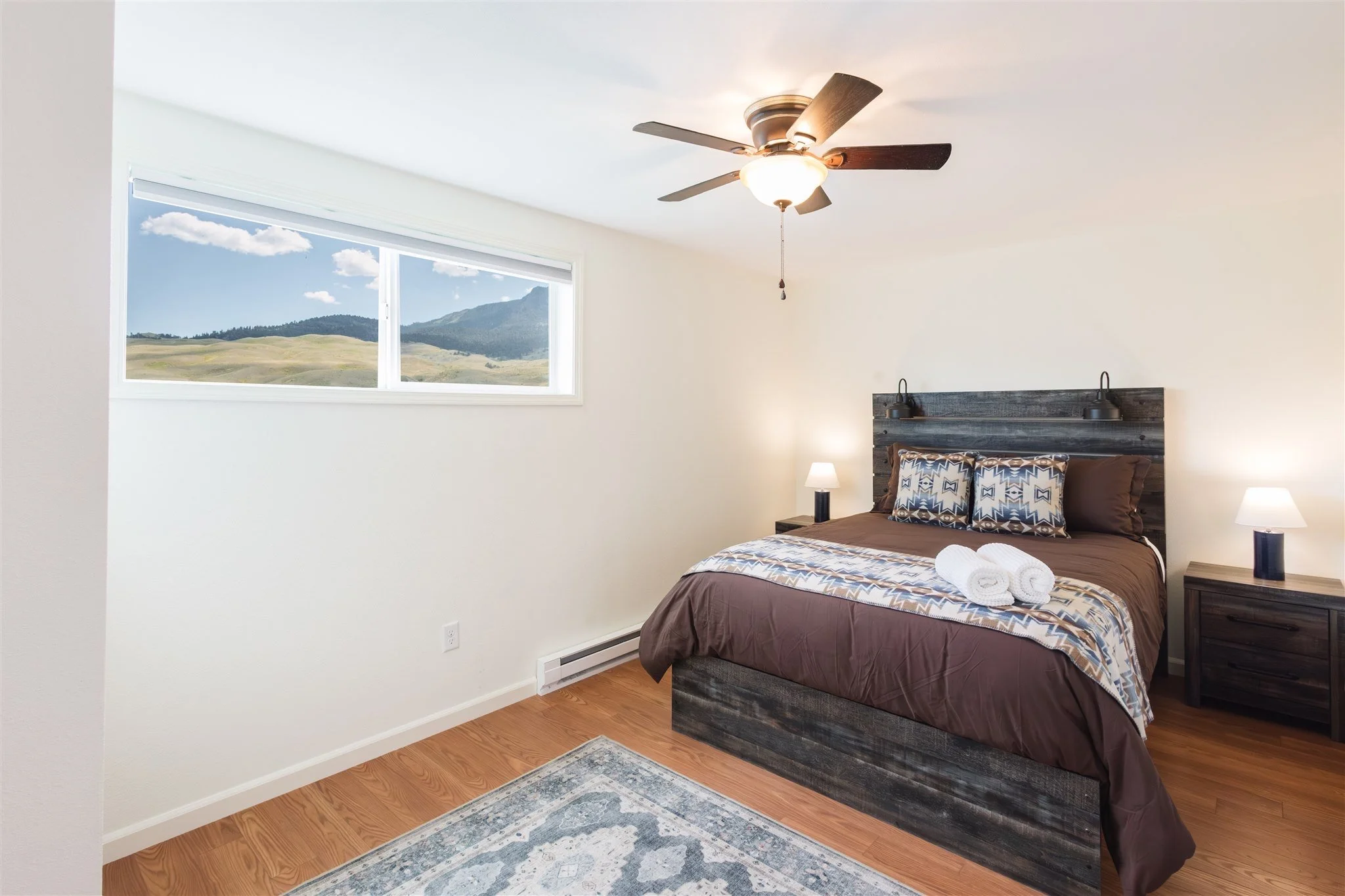 A bedroom with a large window showing a view of hills and mountains, a bed with brown bedding and decorative pillows, two bedside tables with lamps, ceiling fan with light, and a rug on hardwood floor.