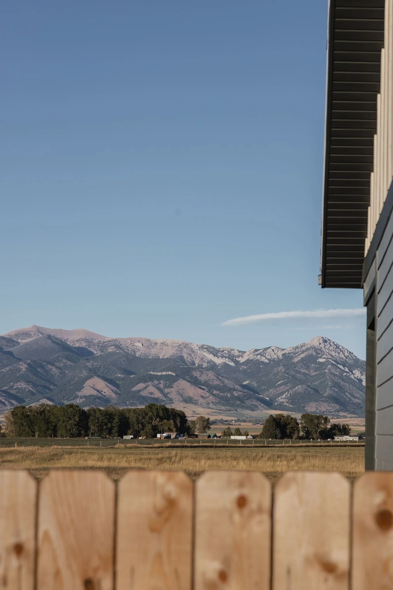 View of mountains and a clear blue sky, seen from behind a wooden fence and part of a building's roof.