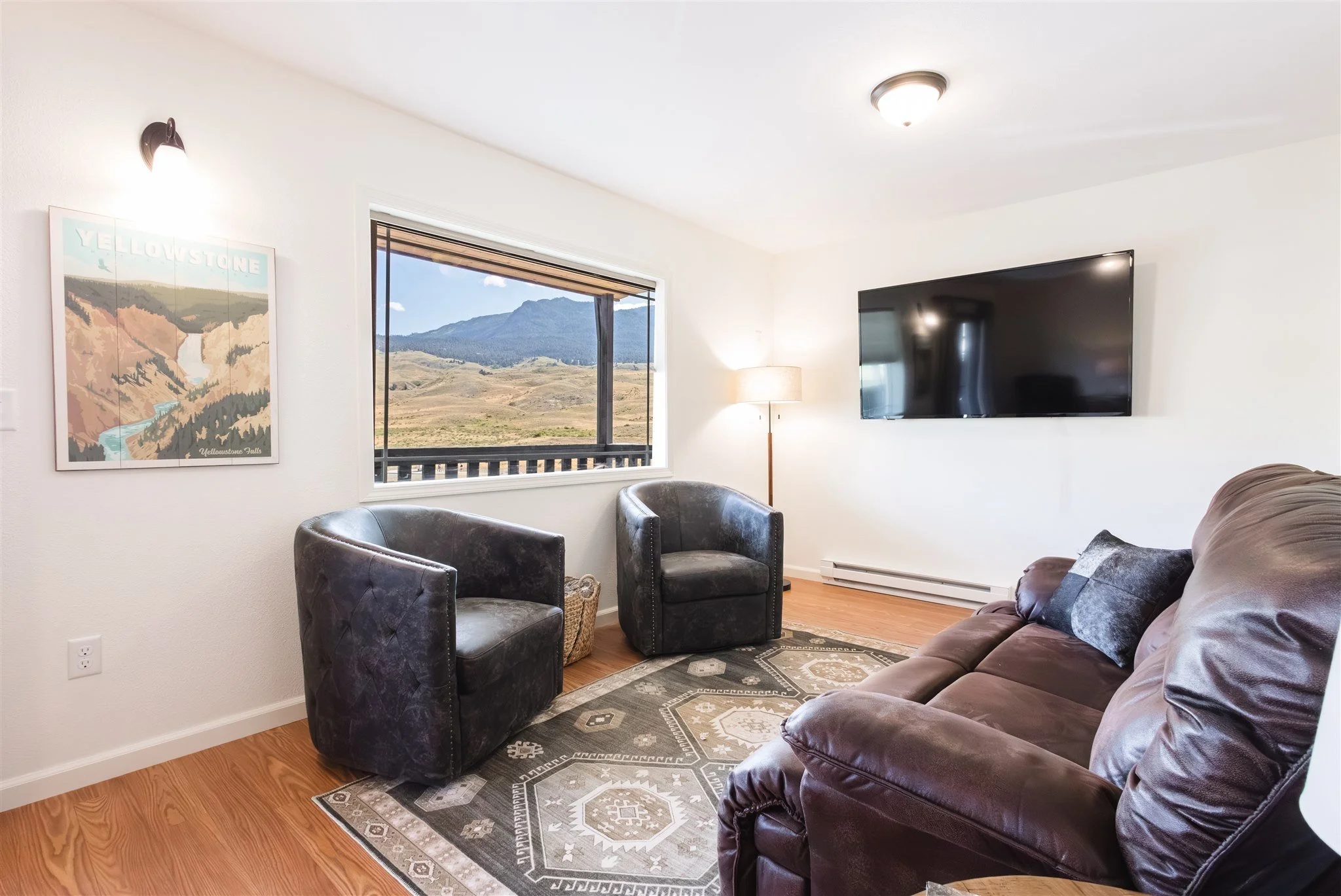 Living room with a large window showcasing a mountain landscape, a wall-mounted TV, a floor lamp, a brown leather sofa, two black armchairs, and a patterned rug.