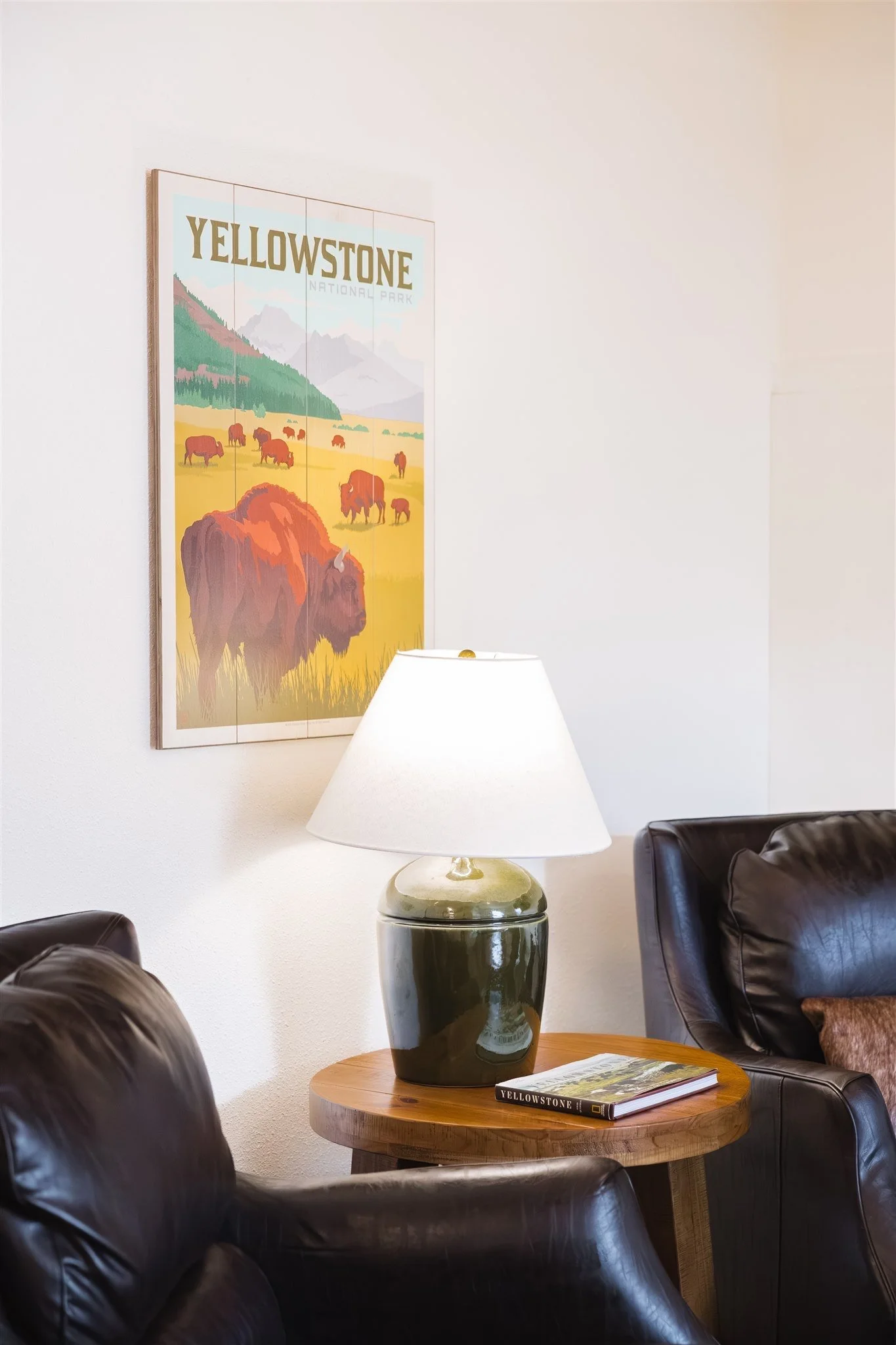 Interior of a lounge or waiting area with a wooden round table holding a green ceramic lamp and a book titled 'Yellowstone.' Behind the table is a black leather armchair, and part of another black leather chair is visible in the foreground. On the wa