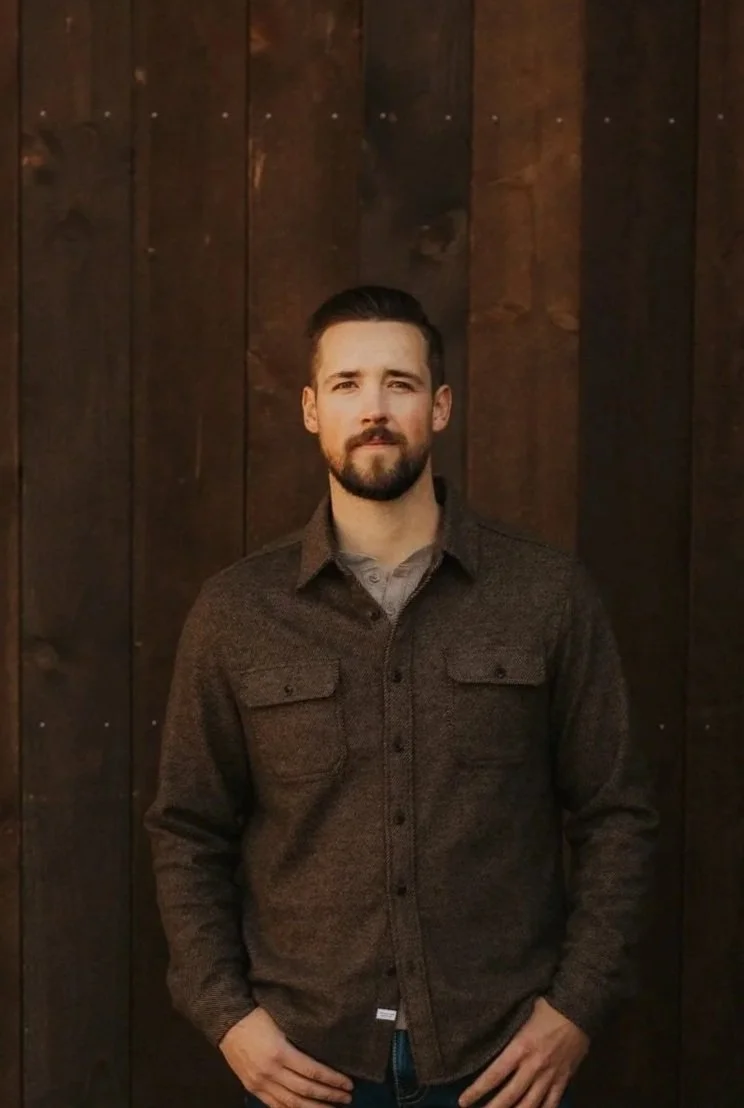 A man with a beard and mustache wearing a black cap and brown jacket, standing in front of snow-capped mountains during sunset or sunrise.