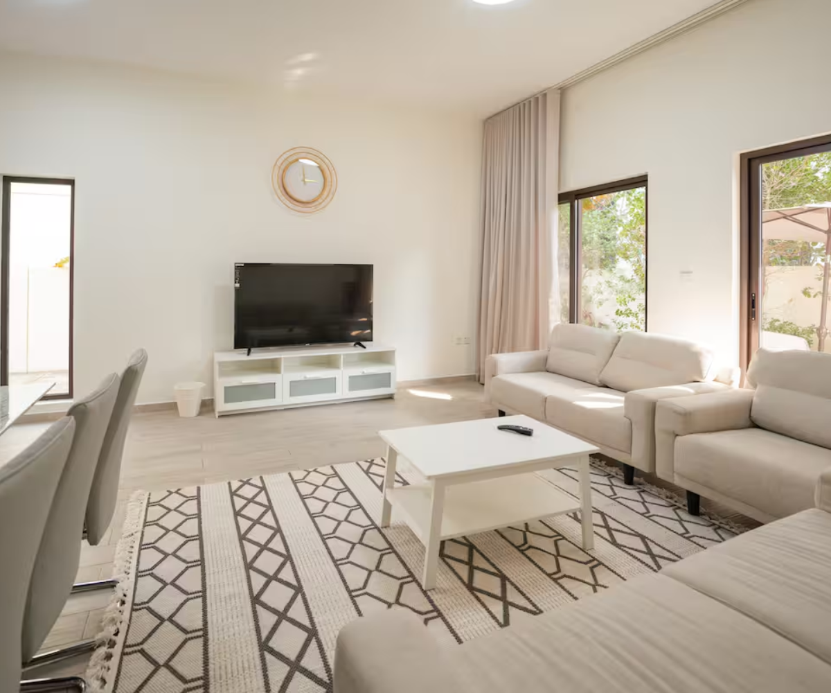 Modern living room with beige sofas, a white coffee table, flat-screen TV on a white console, geometric patterned rug, and wall clock.
