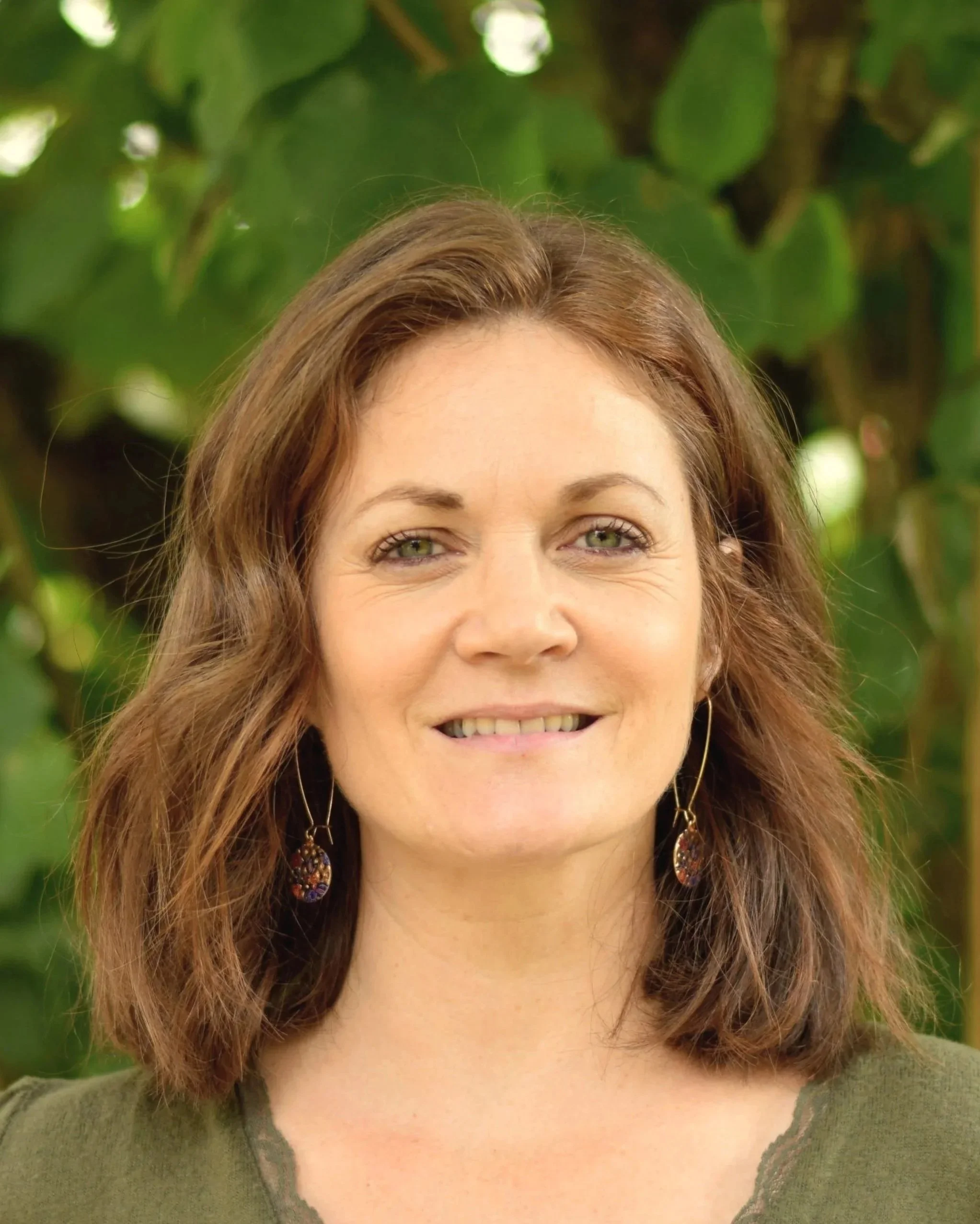 Portrait d'une femme souriante avec des cheveux bruns, portant des boucles d'oreilles colorées, en arrière-plan un feuillage dense.