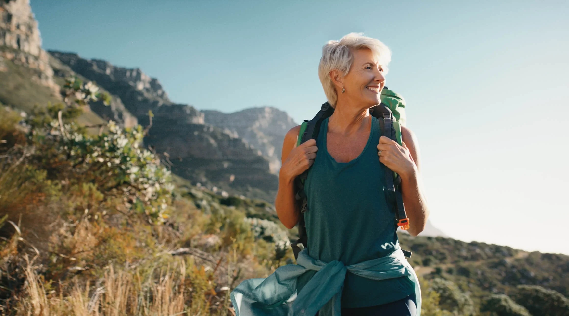 An elderly woman with short gray hair hiking outdoors in a mountainous area, smiling as she looks into the distance, carrying a backpack.