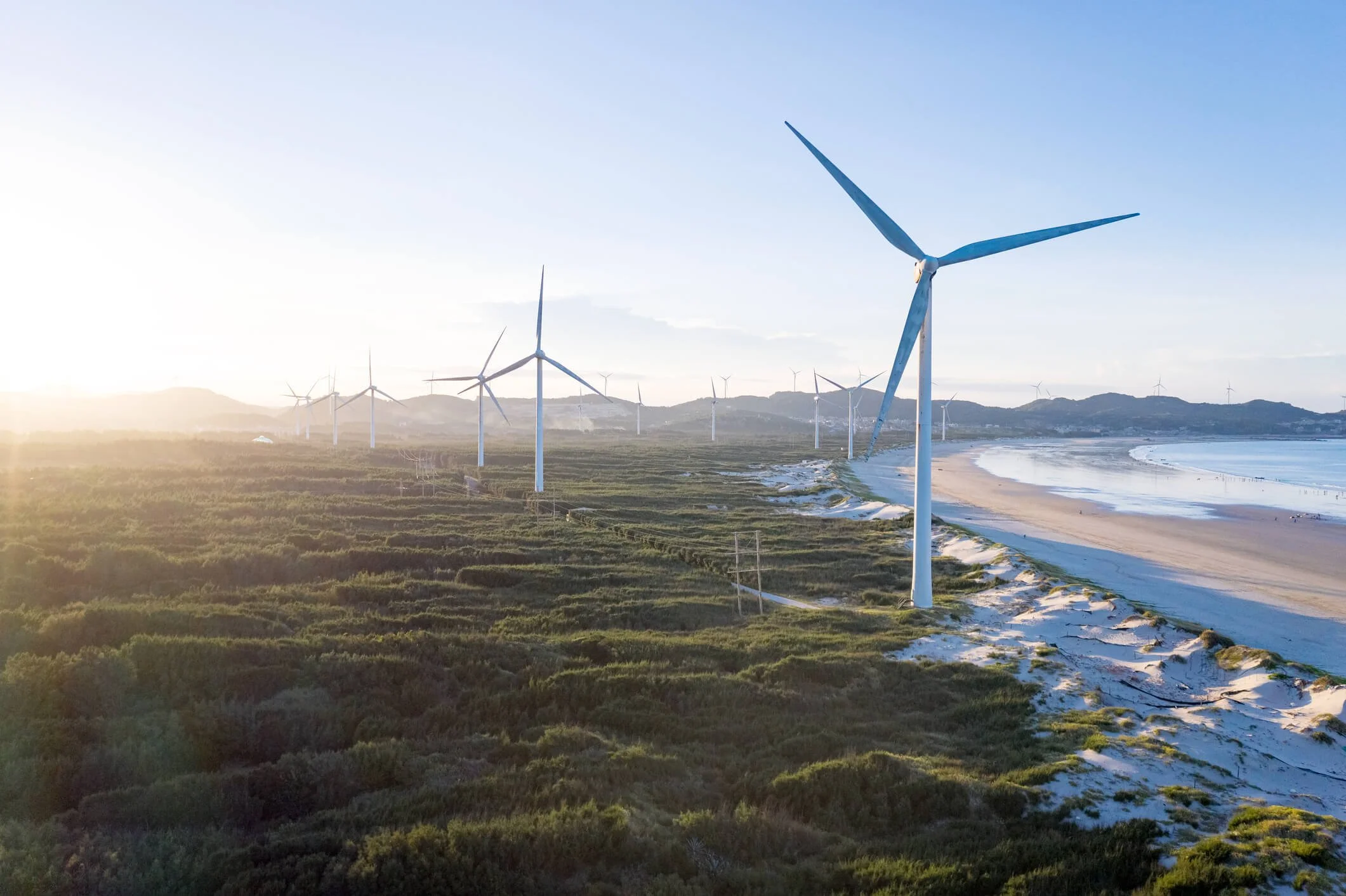 A landscape view of a beach with wind turbines along the shoreline. The scene is illuminated by the setting or rising sun, with hills in the background and a calm ocean on the right.