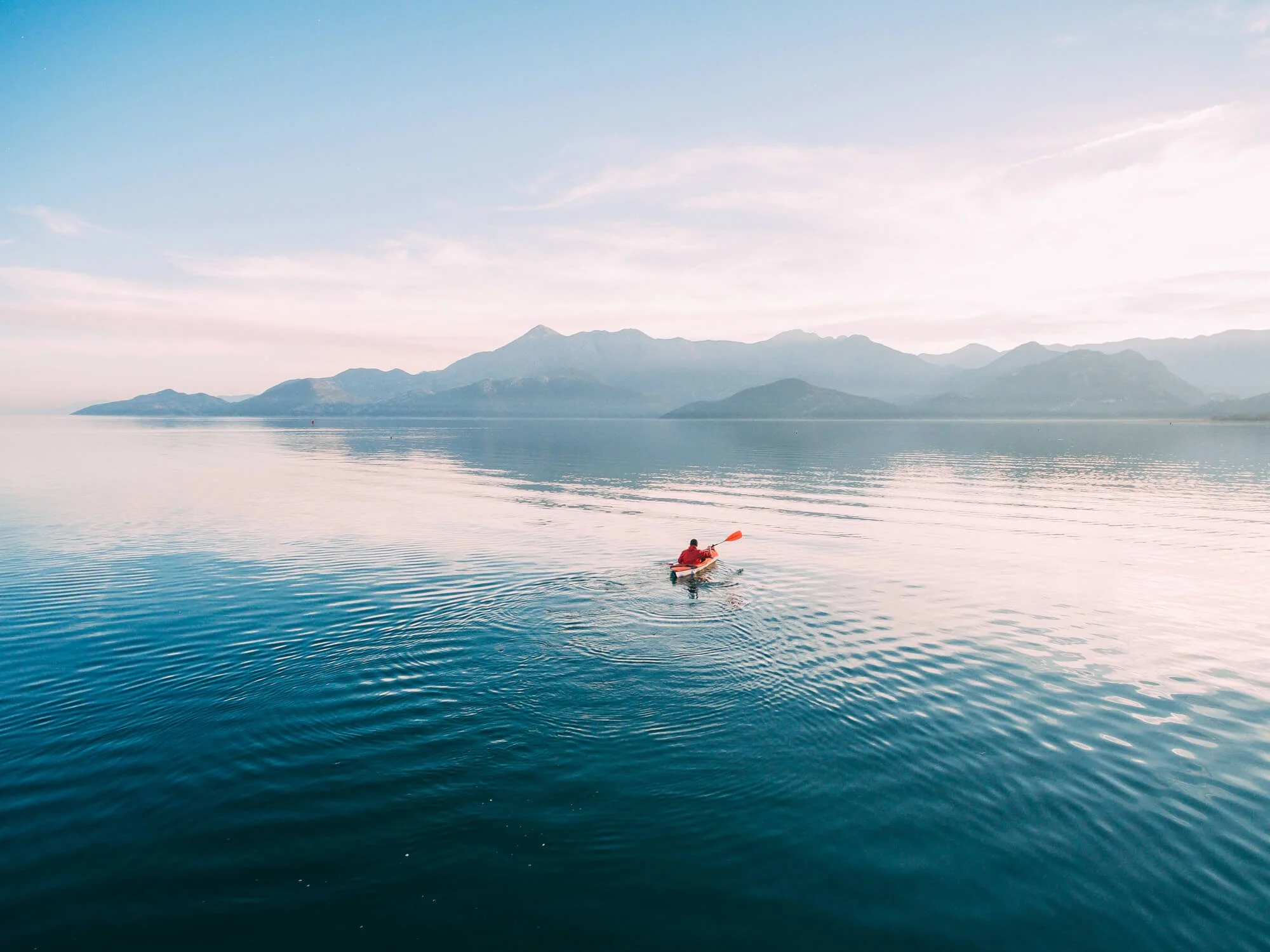 A person in a red jacket kayaking on calm, blue water with mountains in the background and a mostly clear sky.