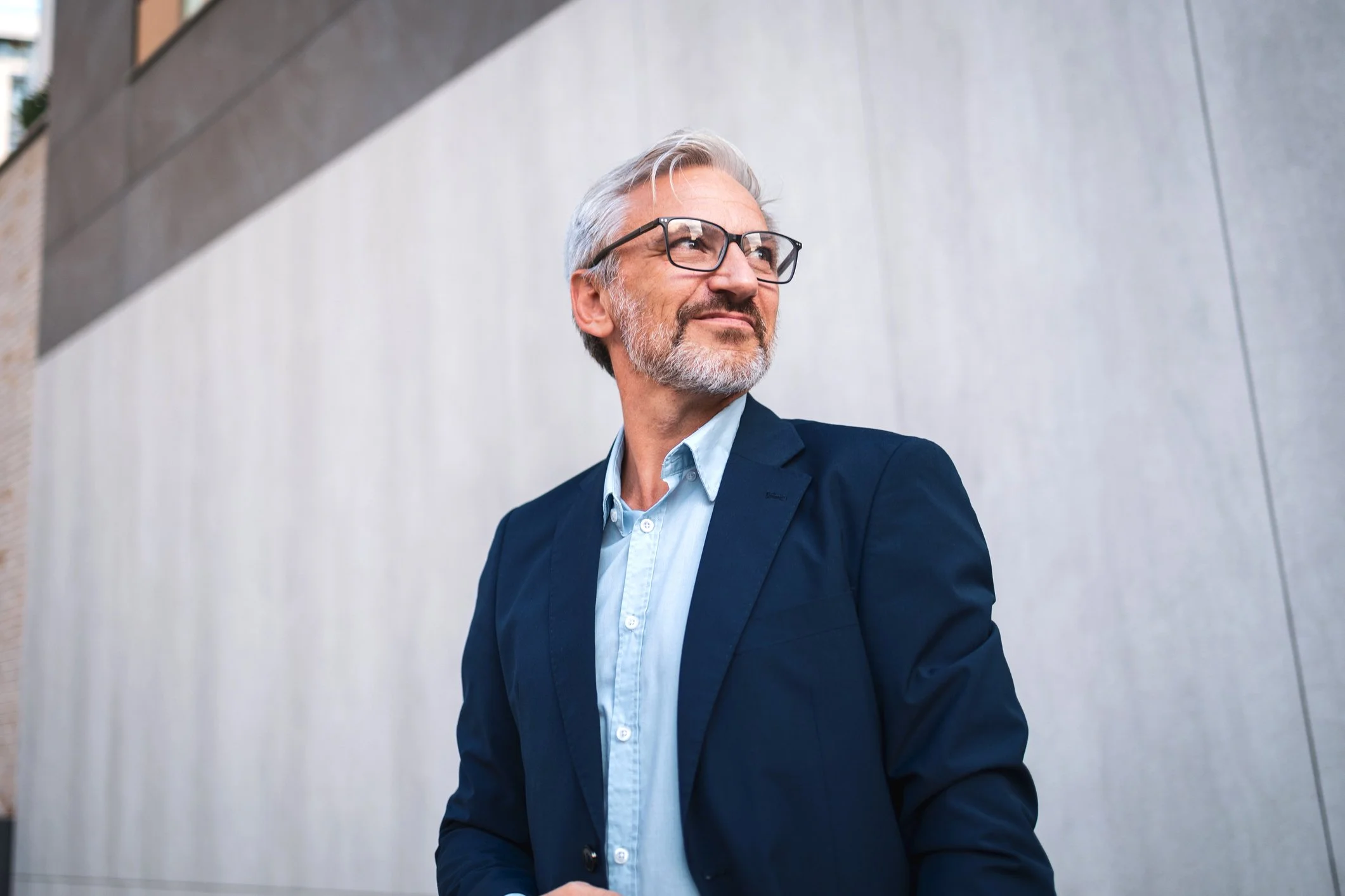 A middle-aged man with gray hair and a beard wearing glasses, a blue blazer, and a light blue shirt, standing against a concrete wall outside, looking to the side with a slight smile.