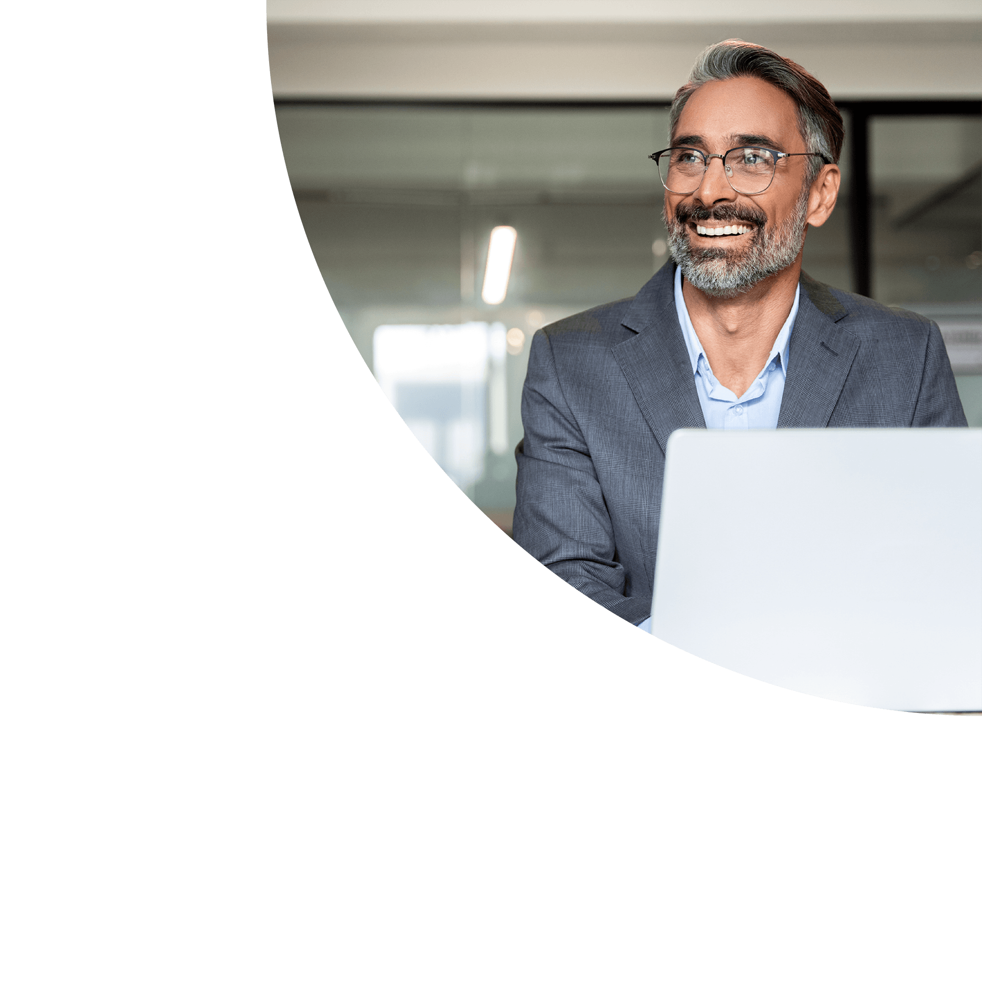 A middle-aged man with gray hair and beard, wearing glasses, in a business suit, smiling while working on a laptop in a modern office.