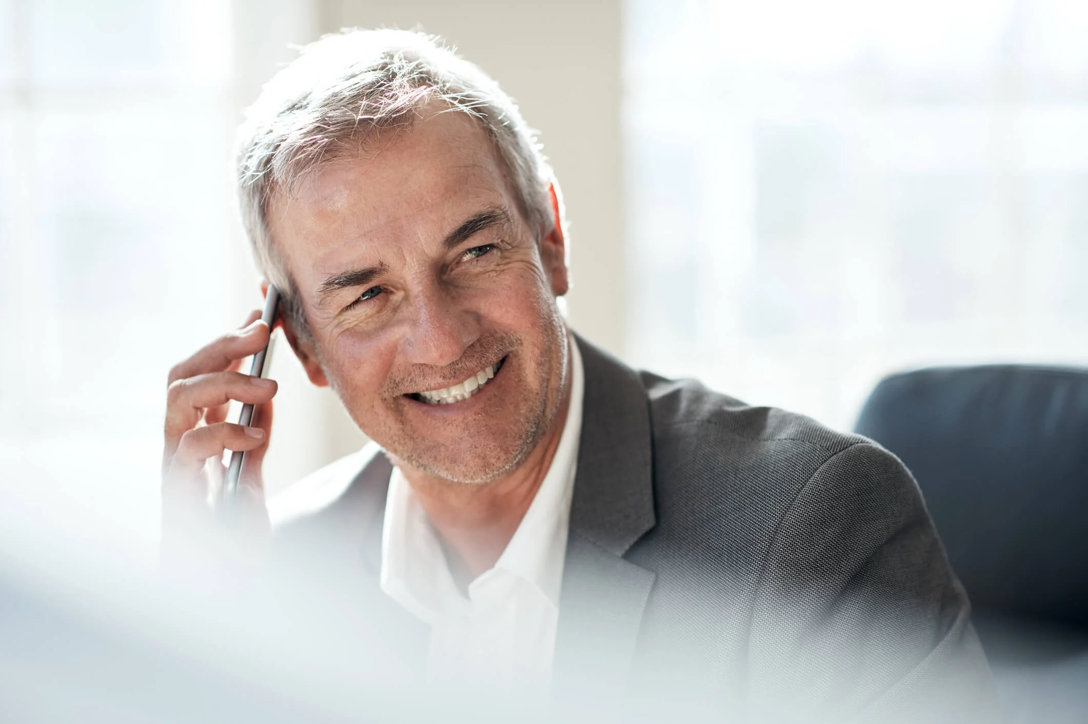 A smiling middle-aged man in a business suit talking on a cellphone in an office with bright natural light.