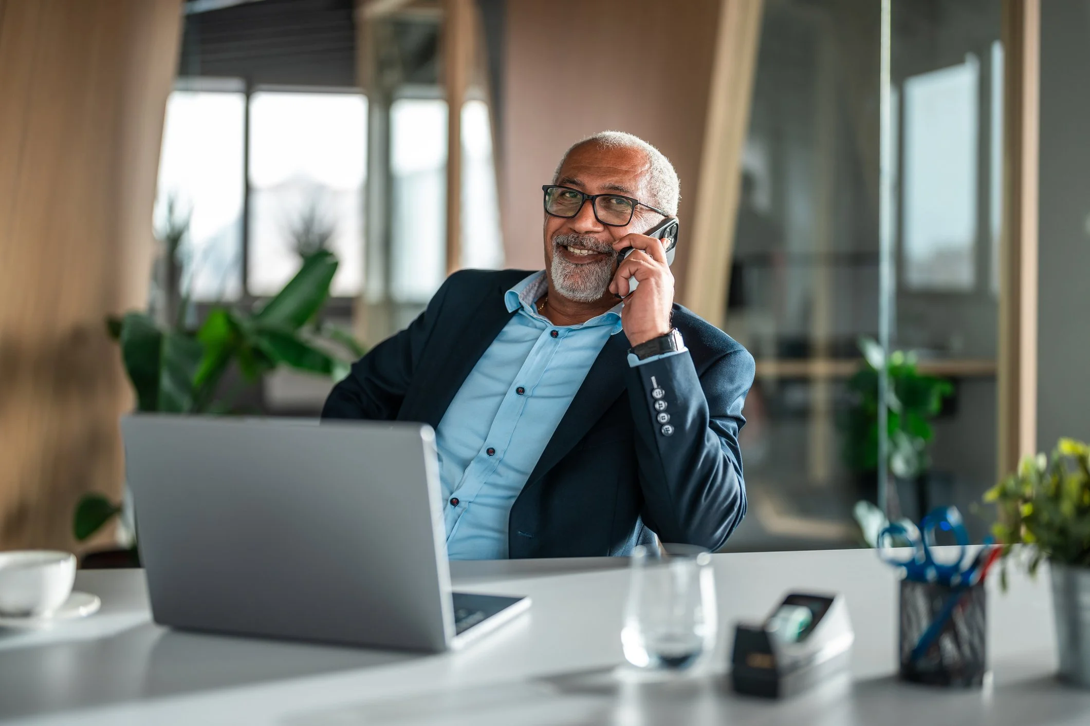 A smiling older man with gray hair and beard, wearing glasses and a suit, talking on a landline phone in an office, with a laptop and office supplies on the desk.