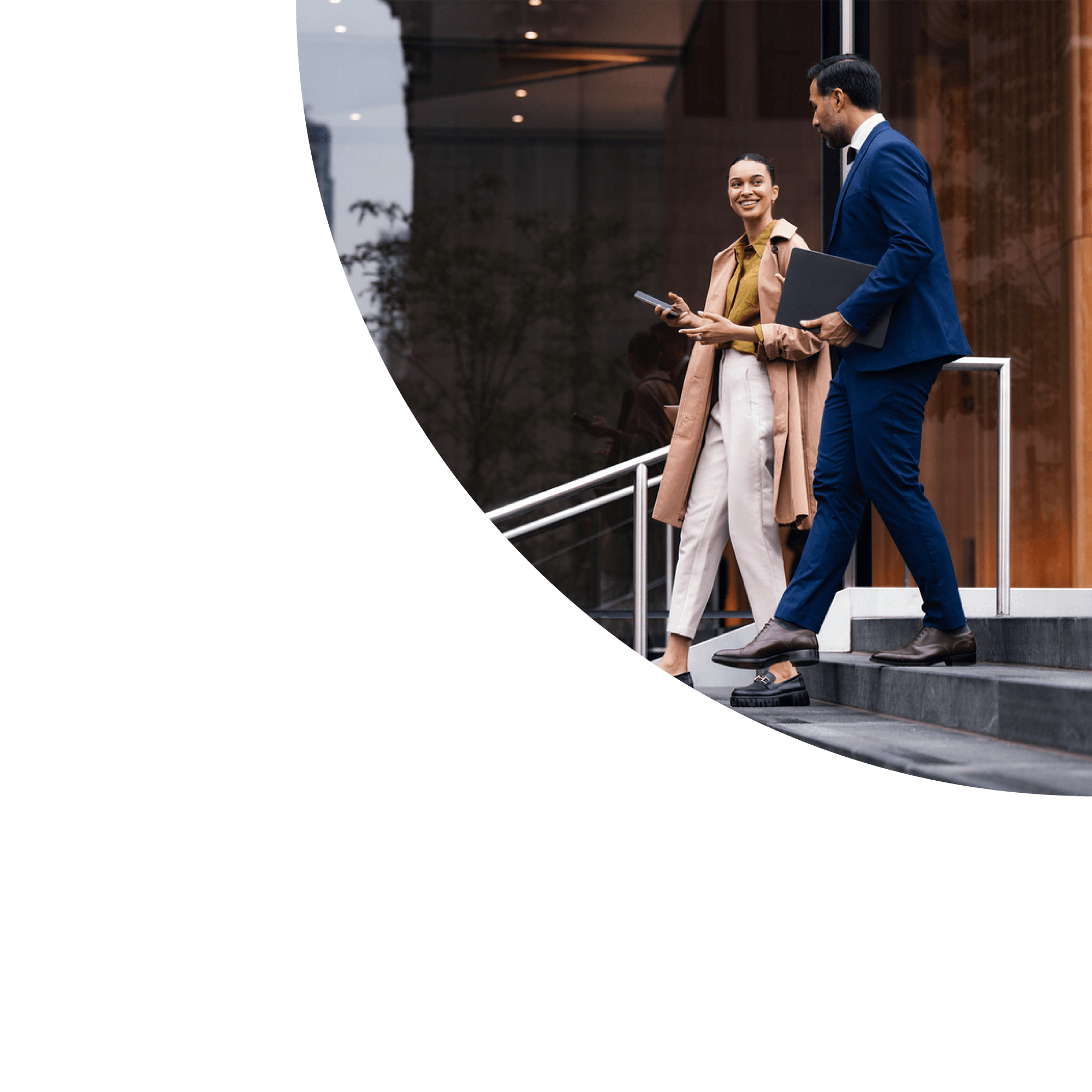 A man in a blue suit and a woman in a beige trench coat walking down outdoor stairs, engaged in conversation, outside a modern building with glass and wood accents.