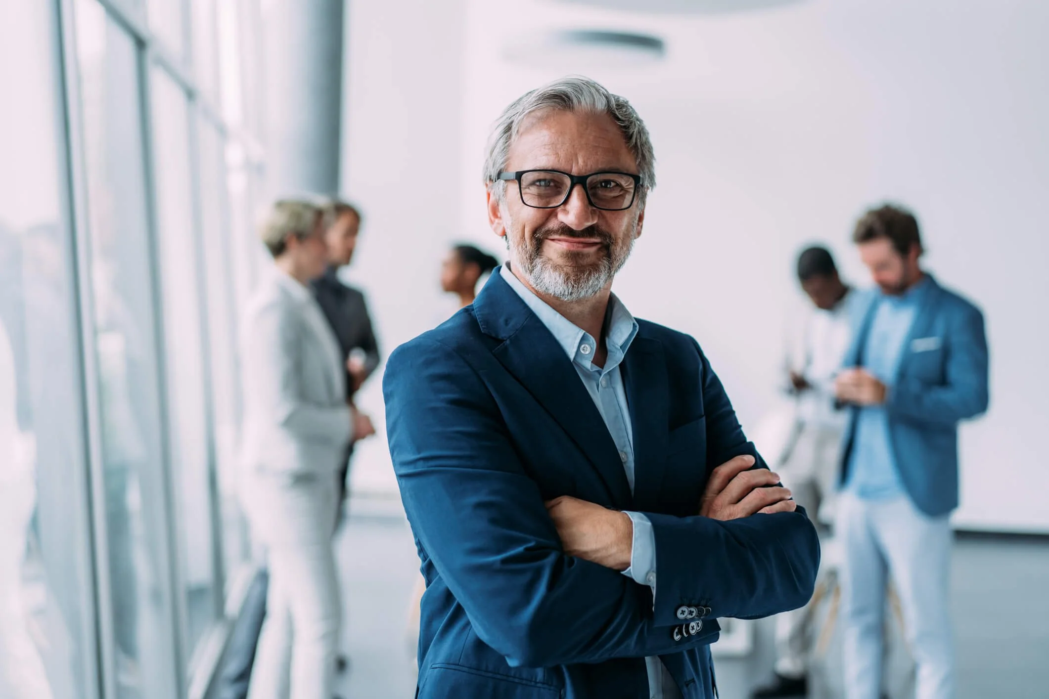 A middle-aged man with gray hair, beard, and glasses standing confidently with arms crossed in a modern office or conference center, with several blurred colleagues in the background.