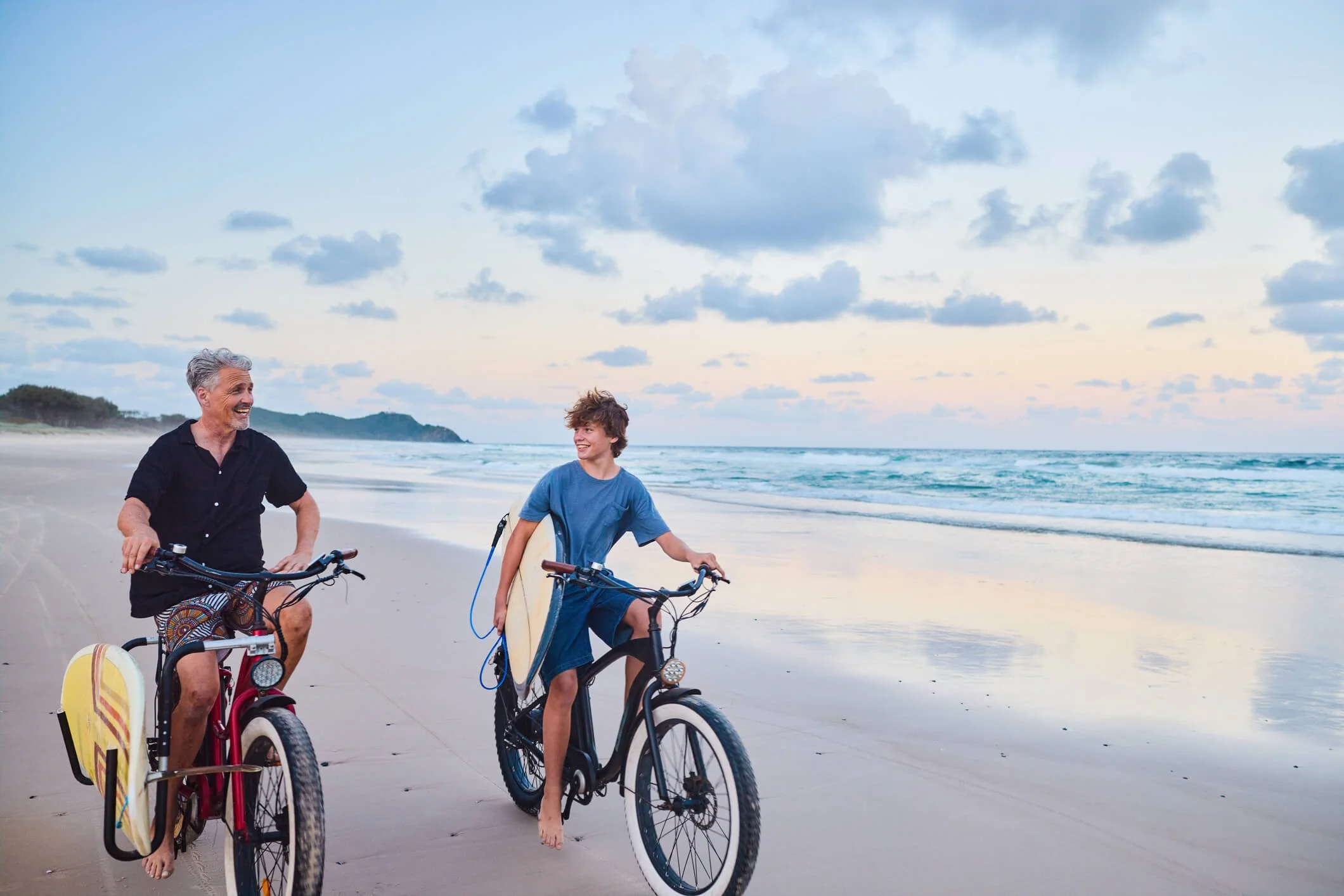 A man and a boy riding bicycles on a beach during sunset, with the man smiling and the boy carrying a surfboard.