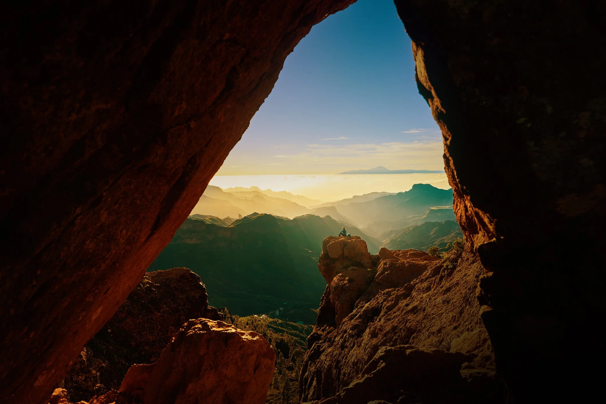 View through a mountain rock opening showing a person sitting on a rock overlooking mountains and a sky with clouds at sunrise or sunset.