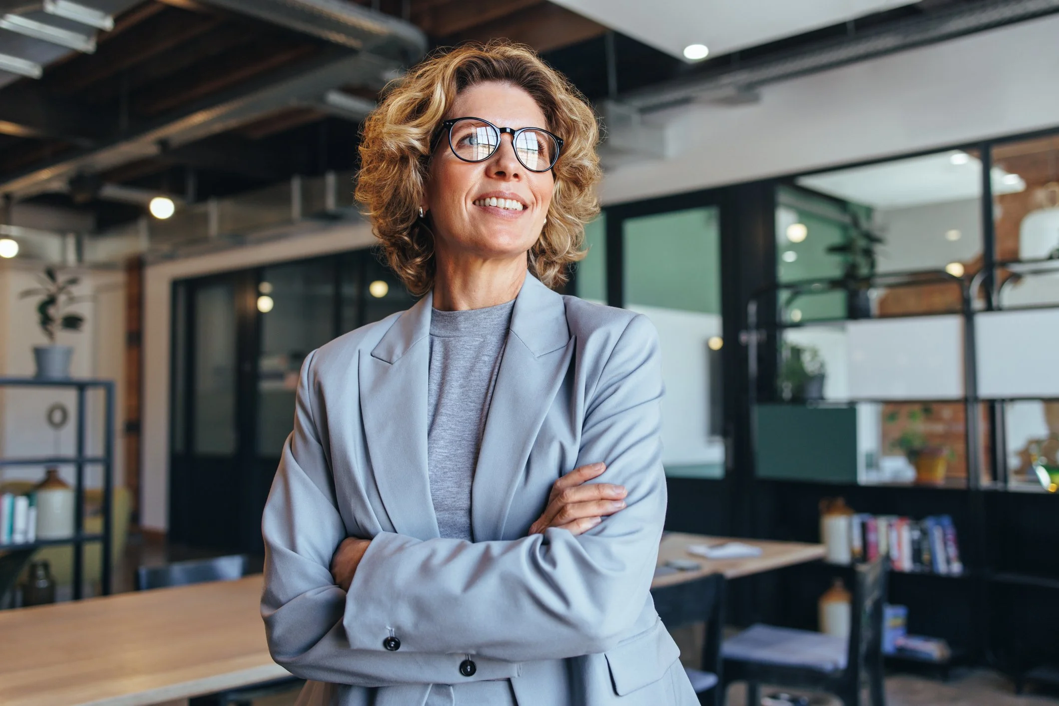 A woman in glasses and a gray blazer stands with crossed arms in an office or coworking space, smiling with a confident expression. The background features a modern interior with shelves, plants, and large windows.