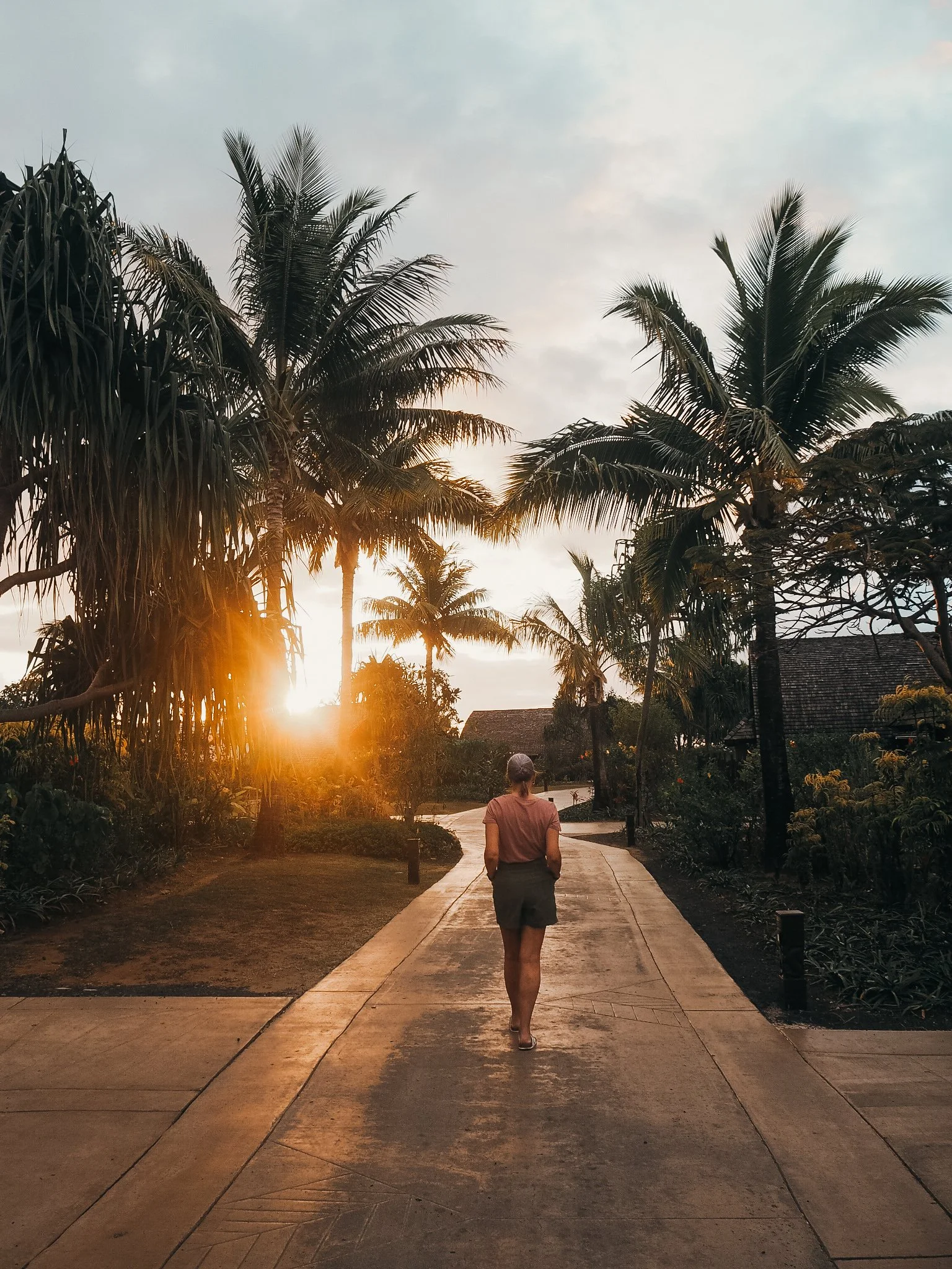 A woman walking on a paved pathway at sunset, surrounded by tall palm trees and tropical vegetation.