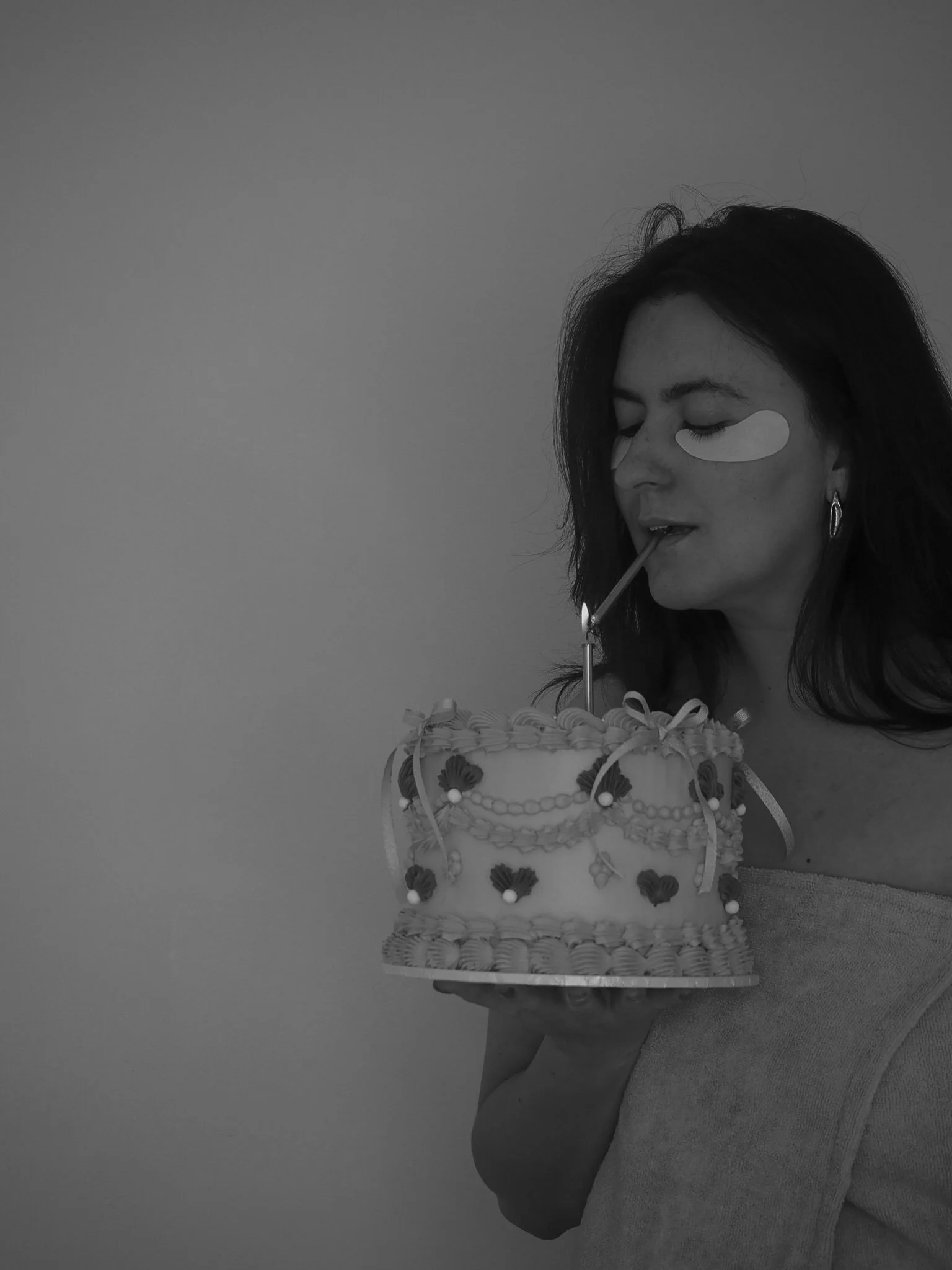 Woman with under-eye patches on face holding a decorated birthday cake with a candle, about to blow out the candle.