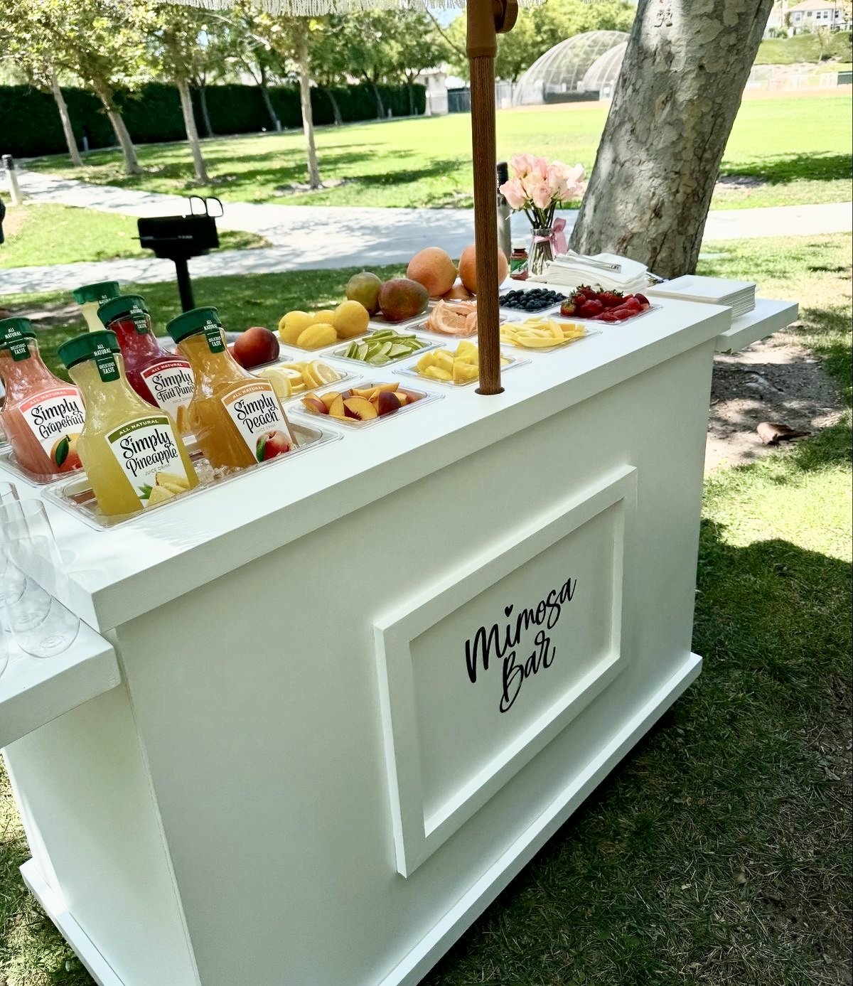 A white bar cart labeled 'Mimosa Bar' set up outdoors with various fruits, juices, and glasses for making mimosas, on a grassy area with trees in the background.